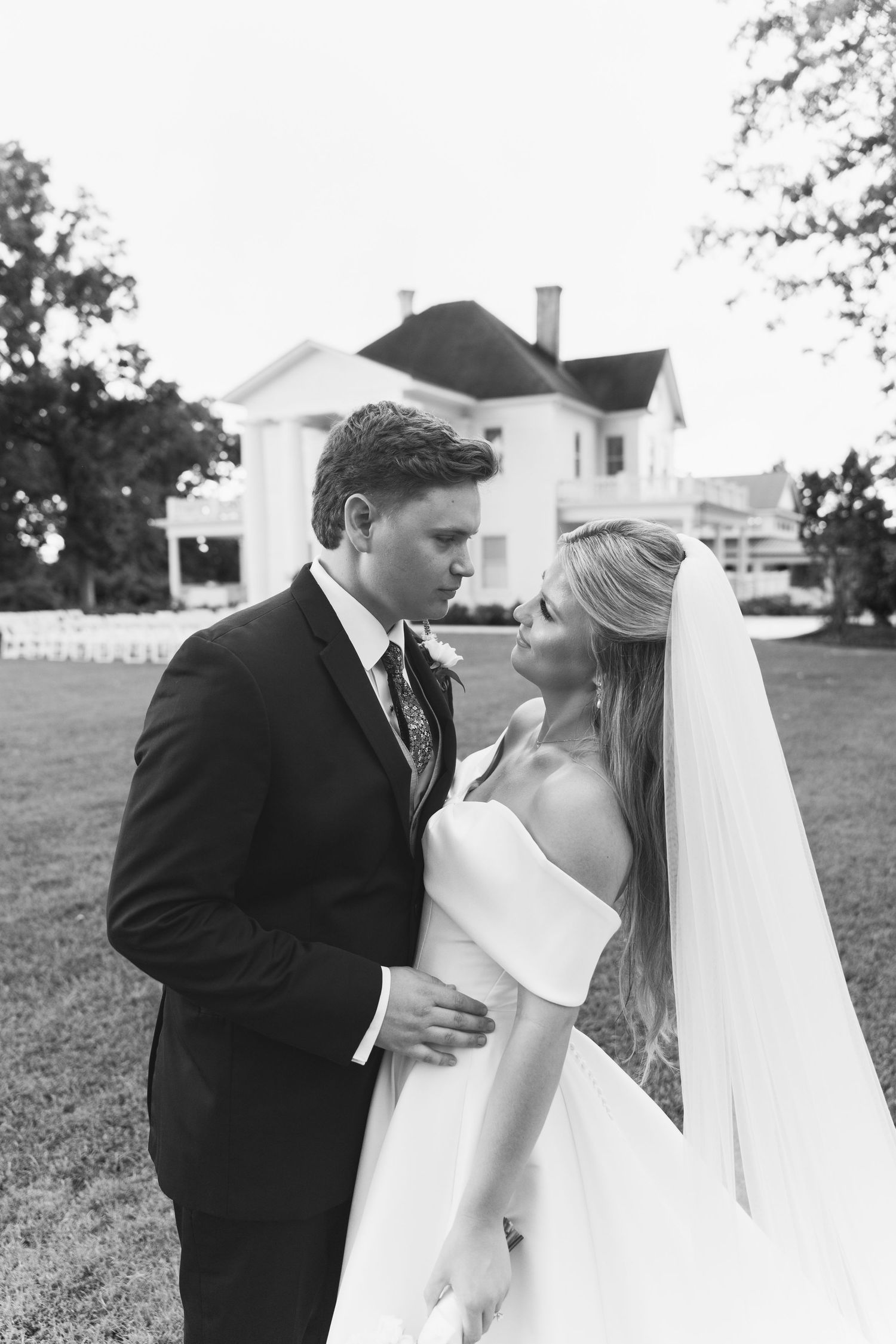 Black and white photo of a bride and groom sharing a romantic moment on a lawn with a white manor house in the background.