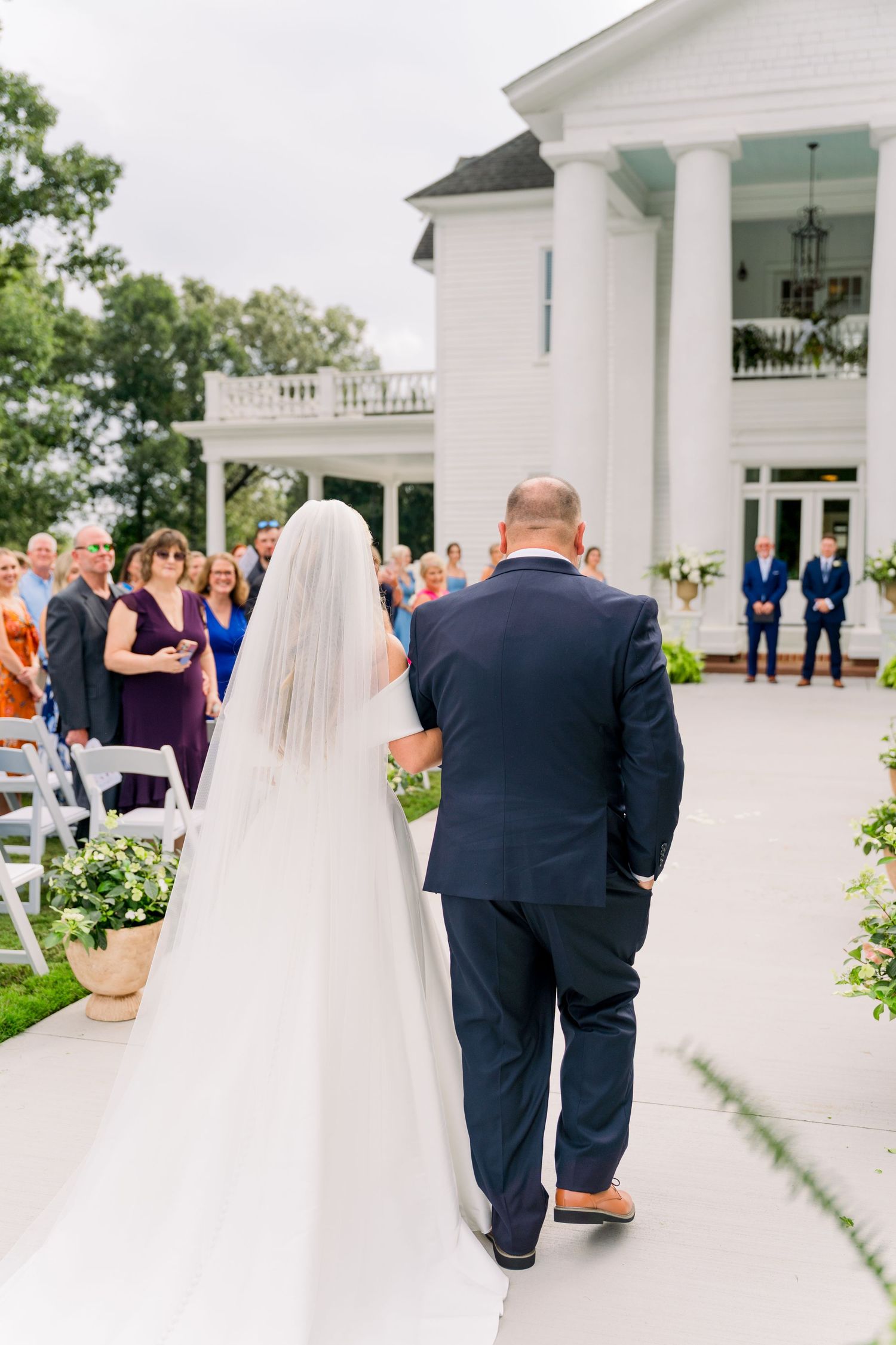 Bride in white gown and veil walks down outdoor aisle with escort toward elegant white mansion wedding venue.