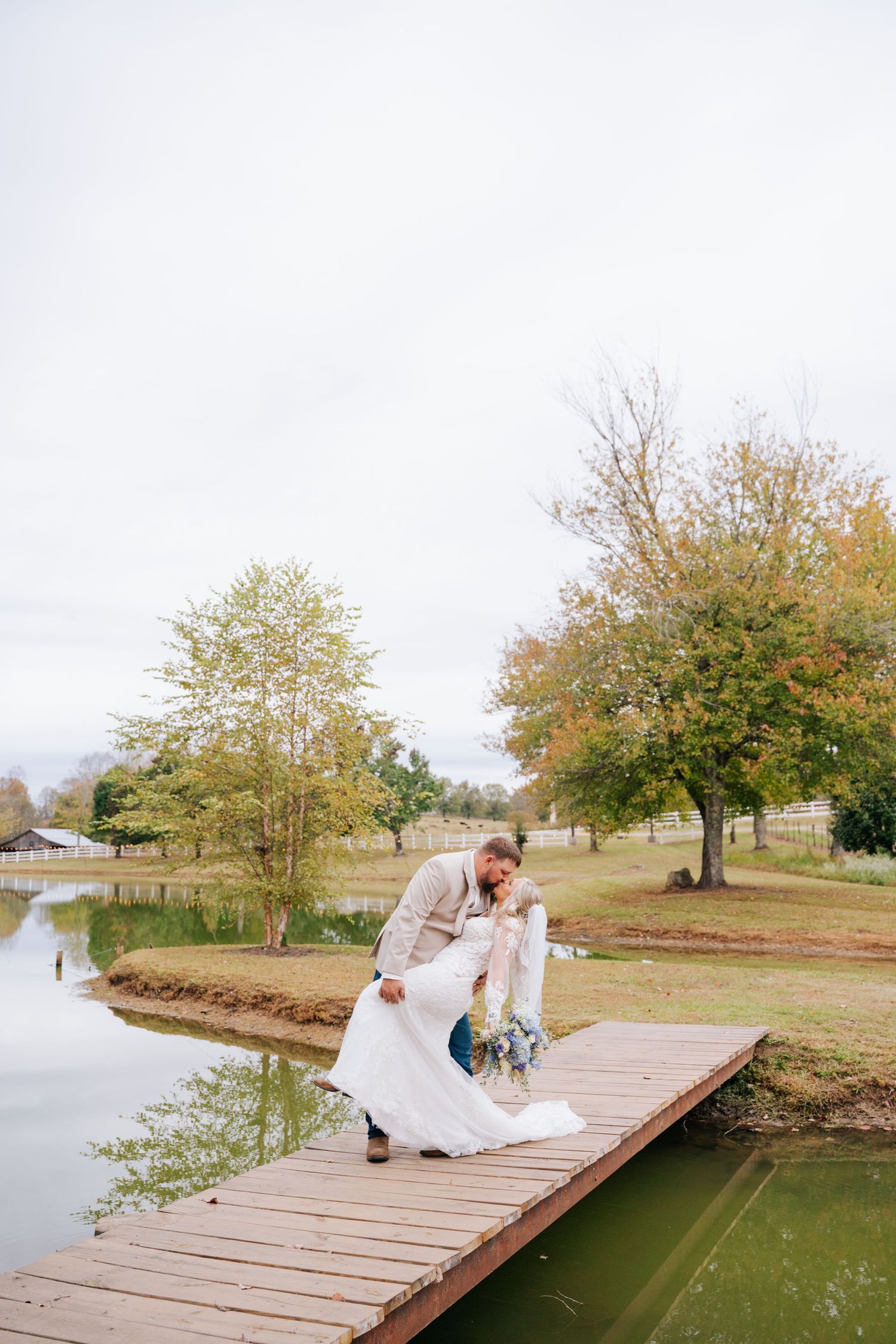 Bride and groom share a romantic dip kiss on a wooden dock beside a serene pond surrounded by autumn trees.