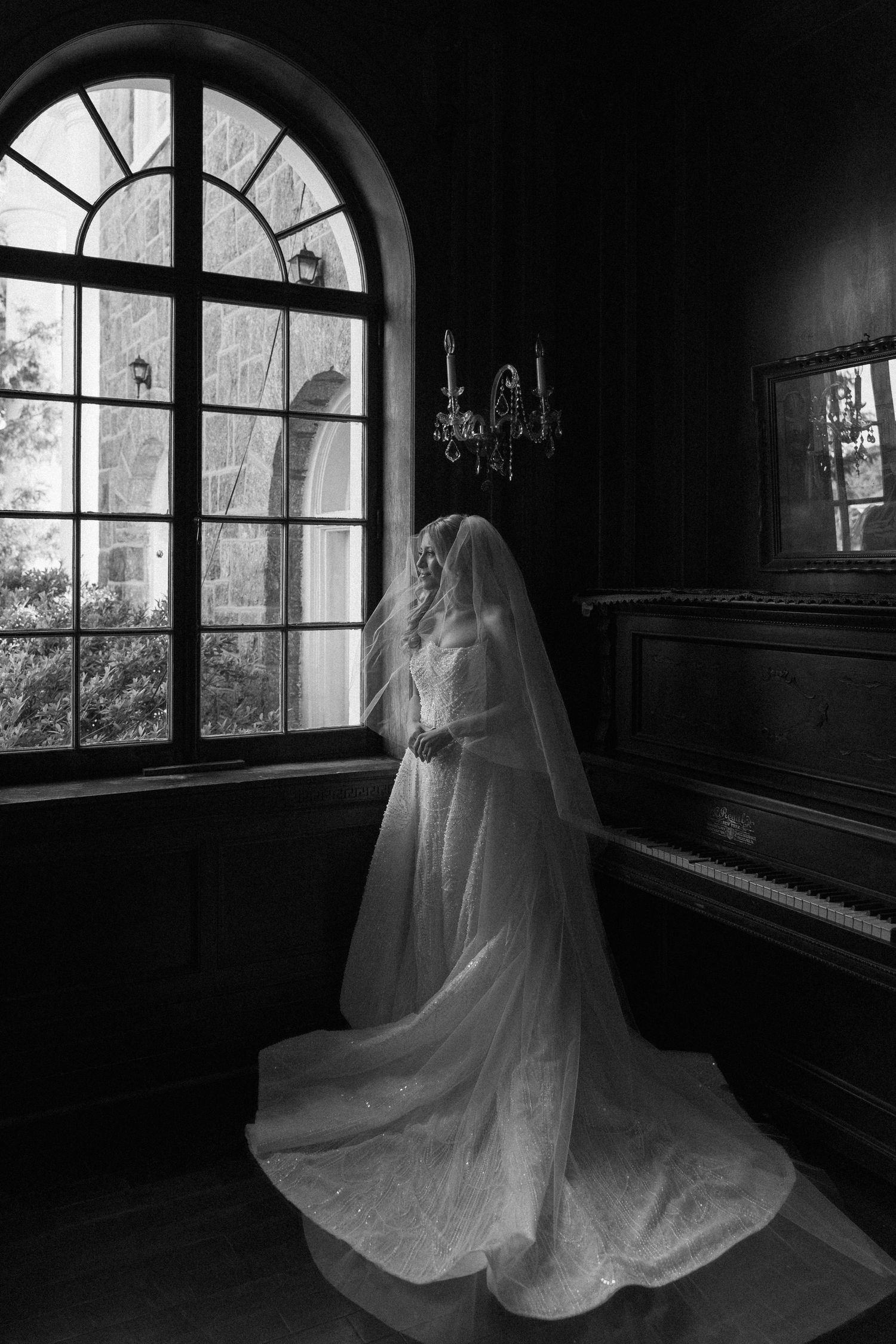 Black and white photo of a bride in a flowing wedding gown and veil standing by an arched window near a piano.