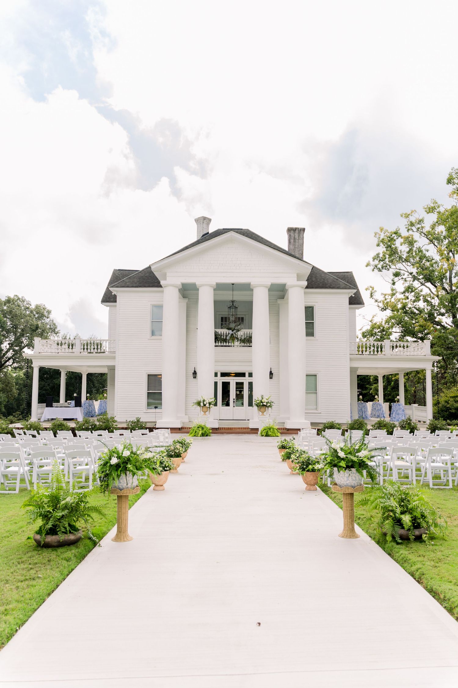 White plantation-style wedding venue with columned facade, lined with ferns and white chairs along a paved aisle.