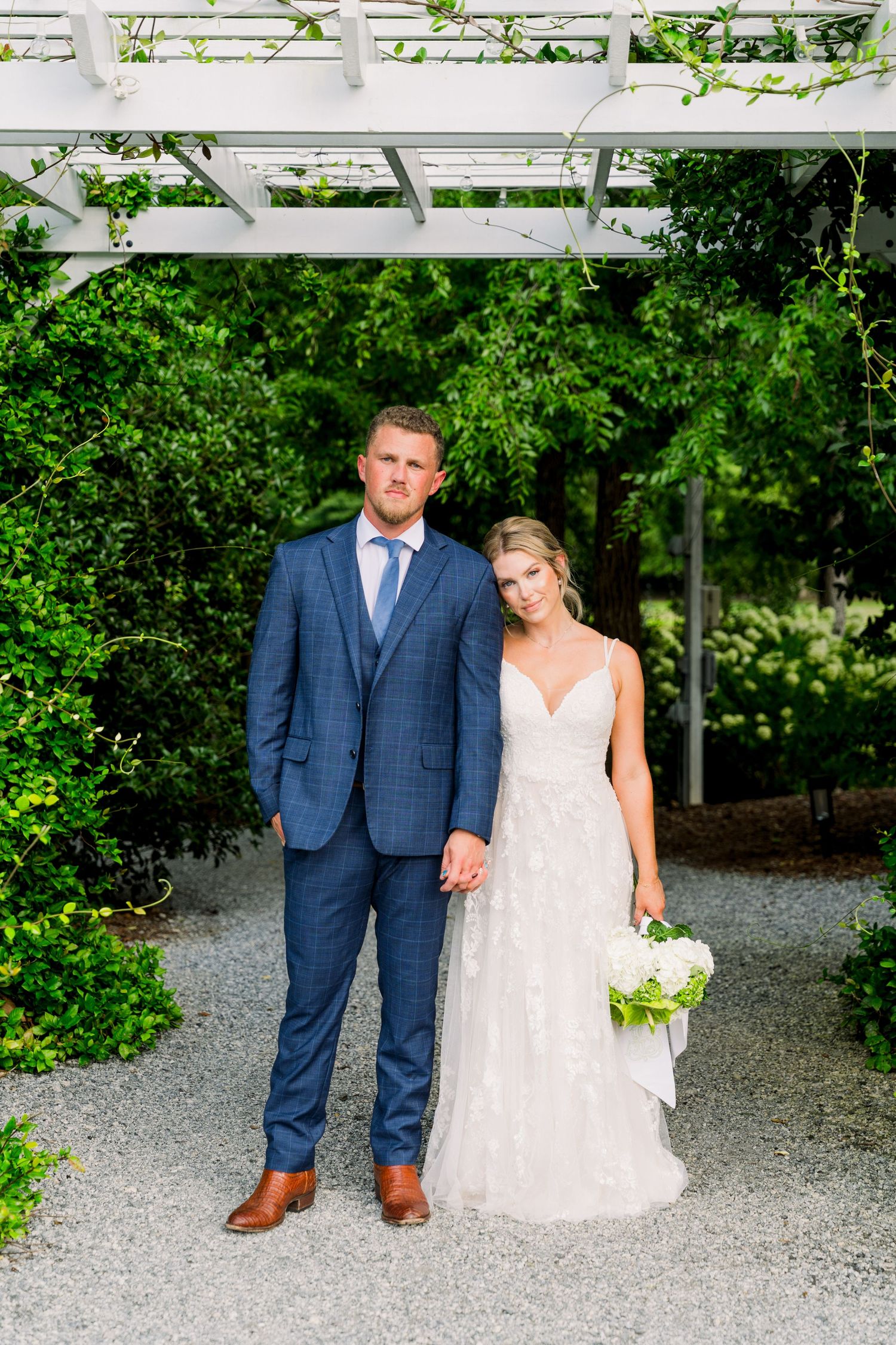 Bride and groom pose under white pergola surrounded by lush greenery, bride holding white bouquet in elegant lace gown.