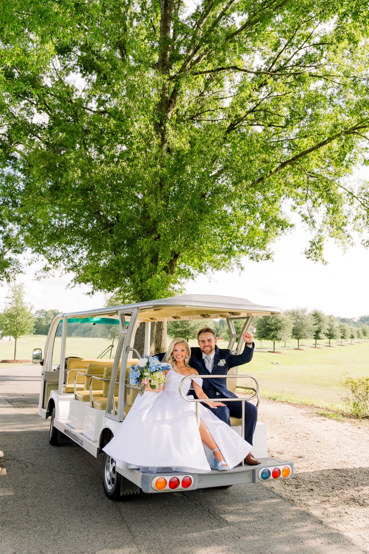 Bride and groom smiling on a white golf cart under a large green tree at an outdoor wedding venue.