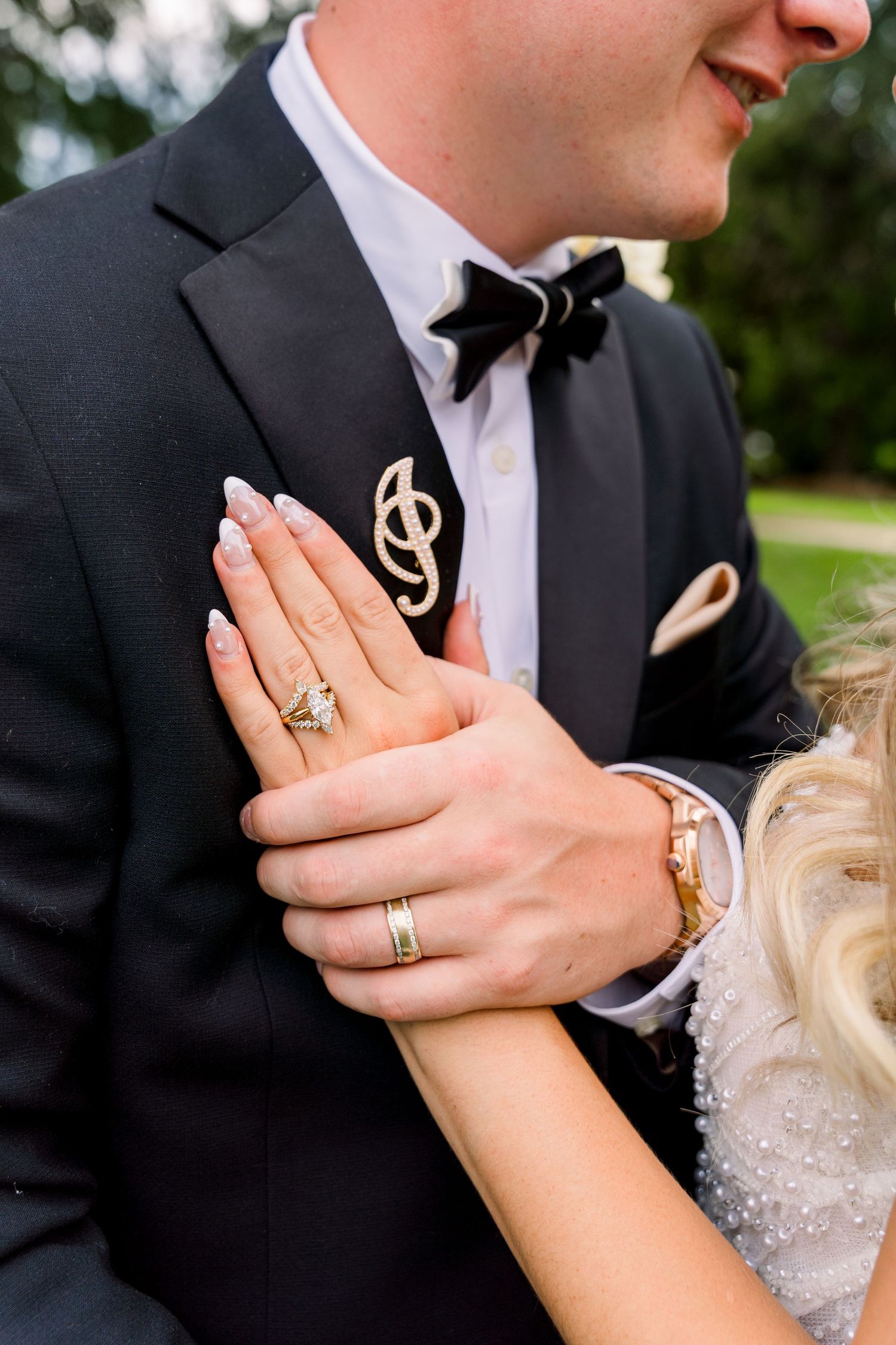 Close-up of wedding couple's hands, bride wearing diamond ring and gold watch, groom in black tuxedo with gold lapel pin.