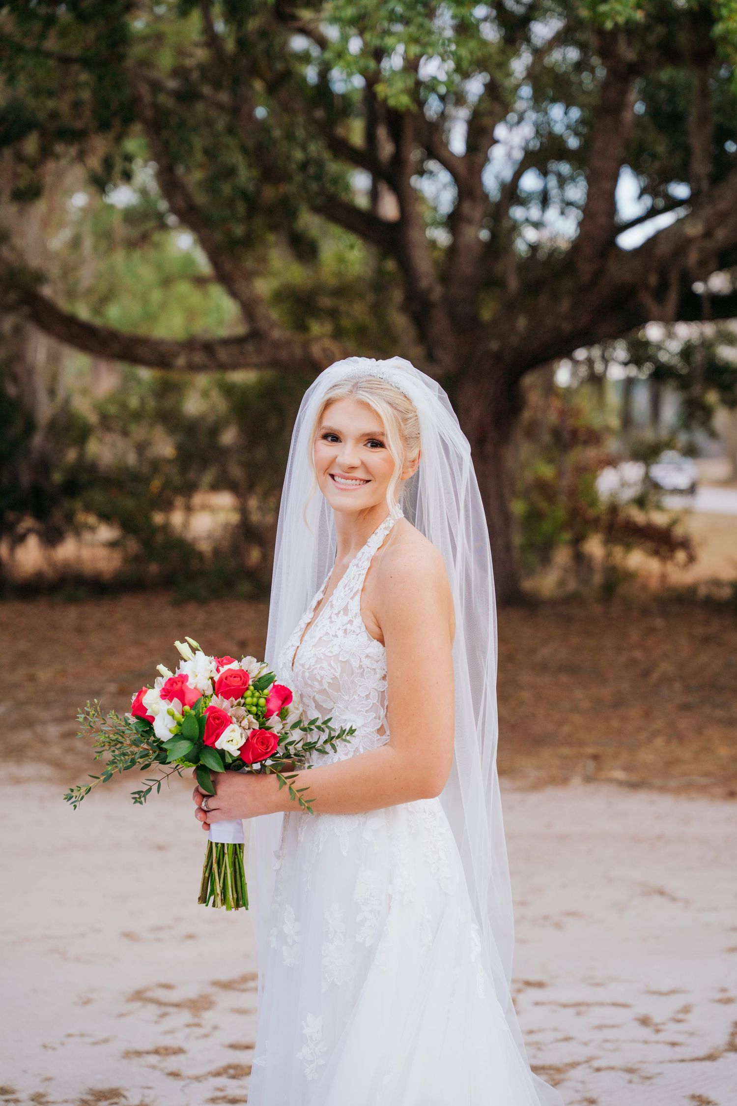A bride in a white lace wedding dress holds a vibrant pink and red bouquet while posing outdoors among pine trees.