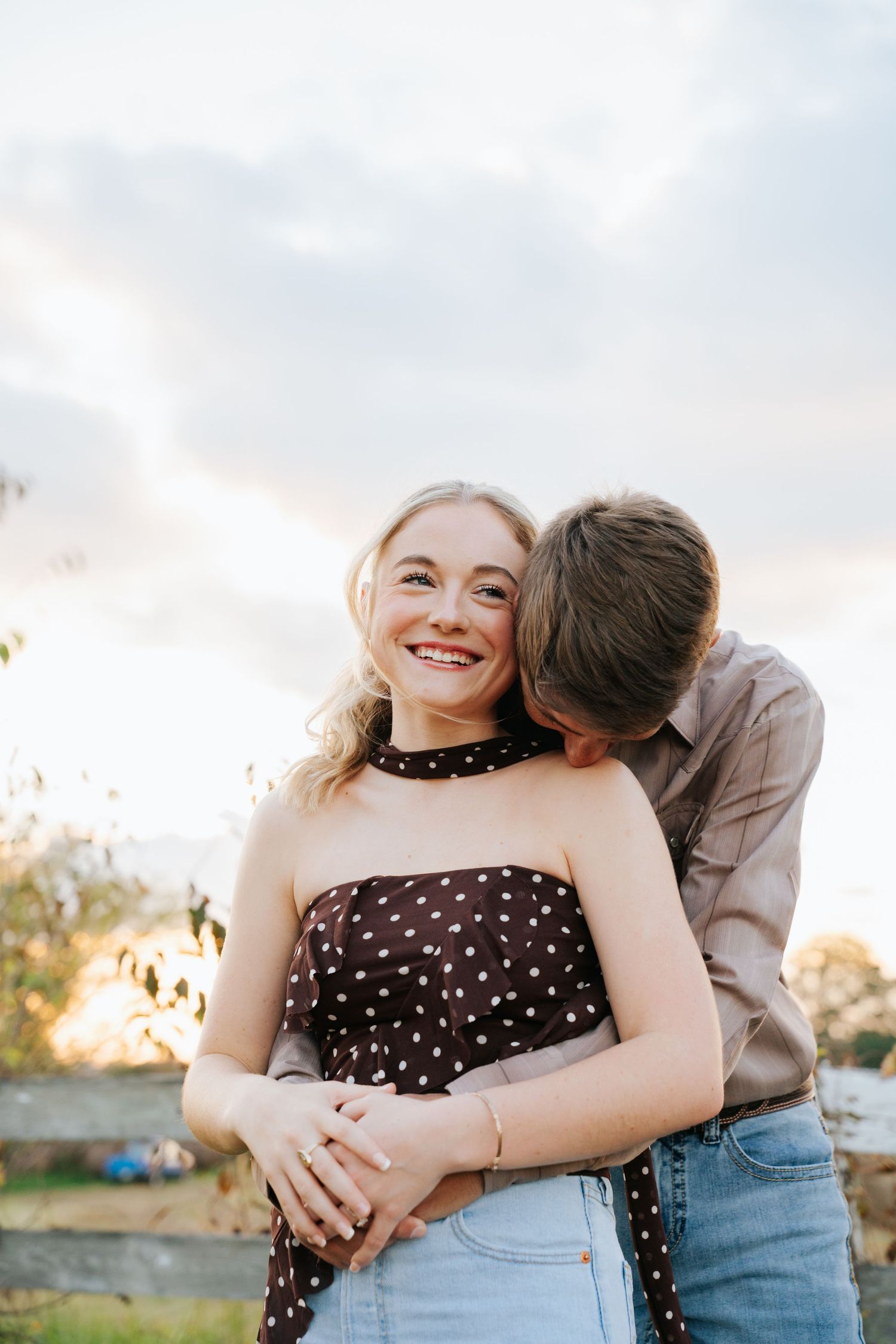 Romantic couple embraces outdoors at sunset, with person in polka dot dress smiling brightly.