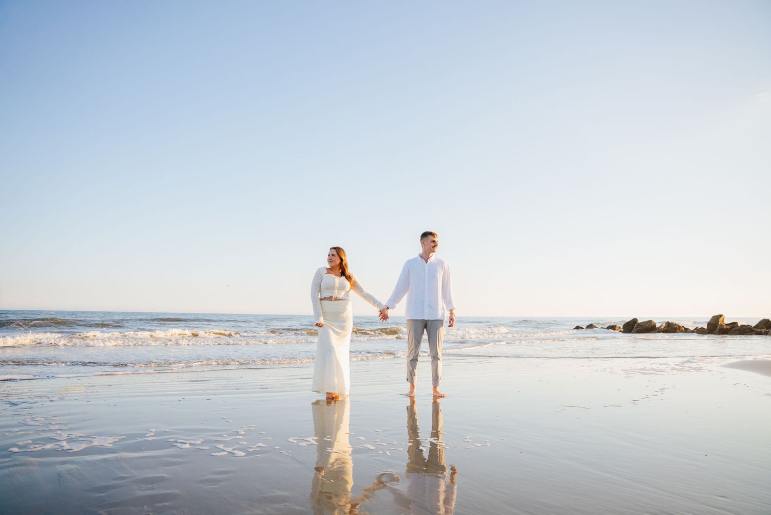 A couple walks hand in hand along a serene beach at sunset with soft waves rolling onto the shore.