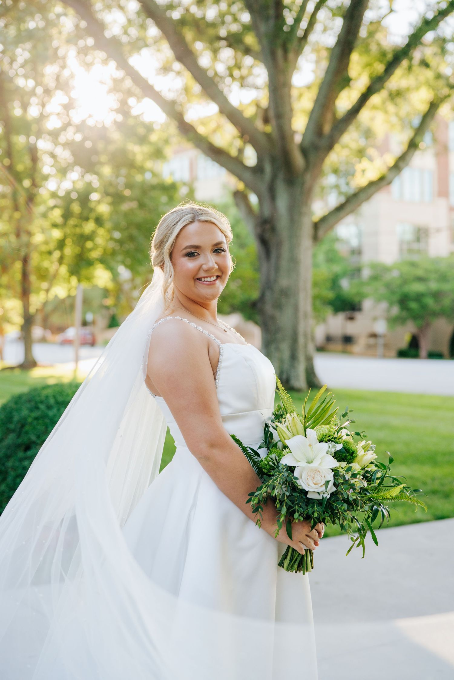 Elegant bridal portrait with flowing train and bouquet against tree-lined backdrop.