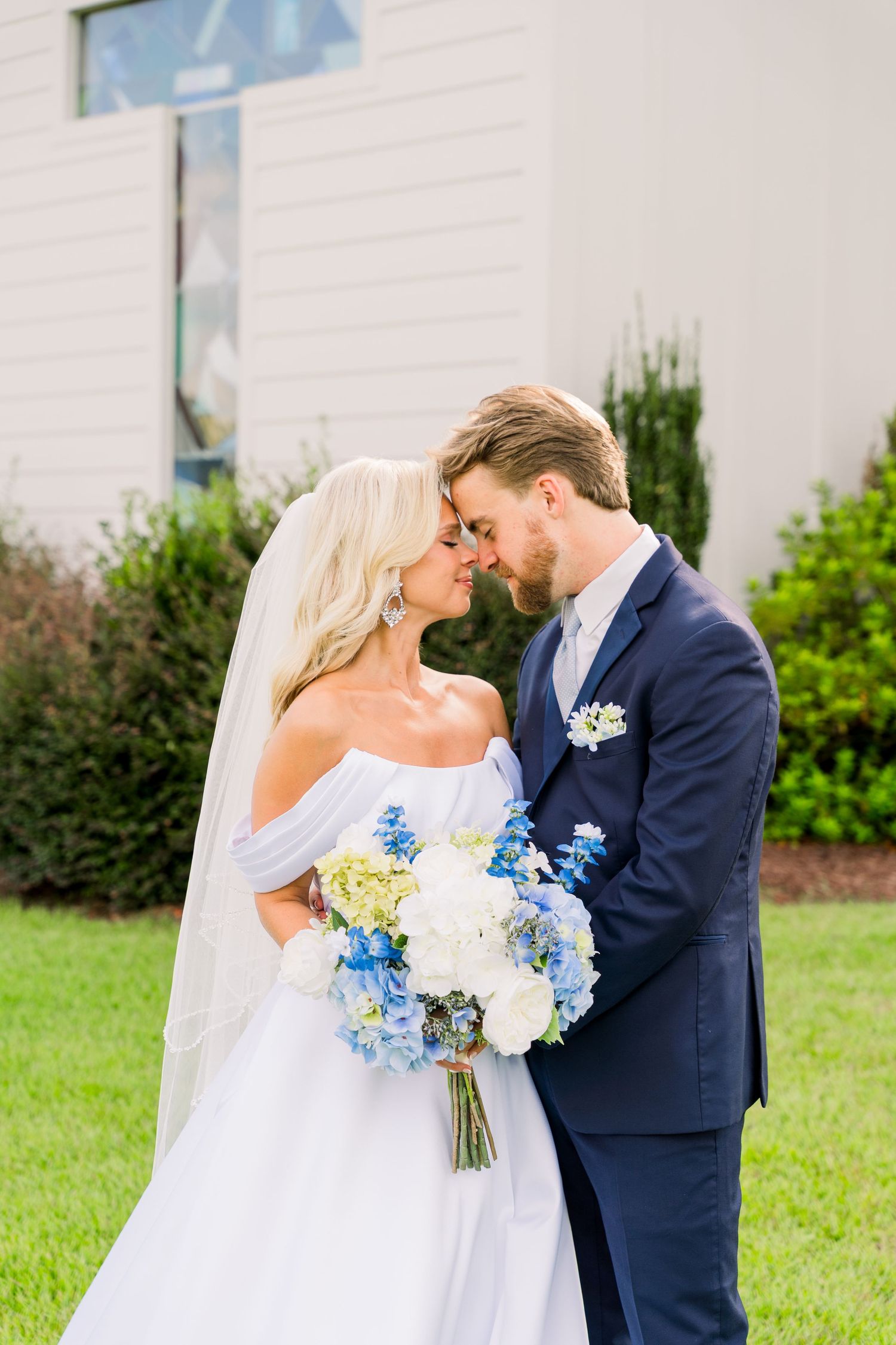 Bride in off-shoulder gown and groom in navy suit share a romantic moment holding blue and white bouquet.