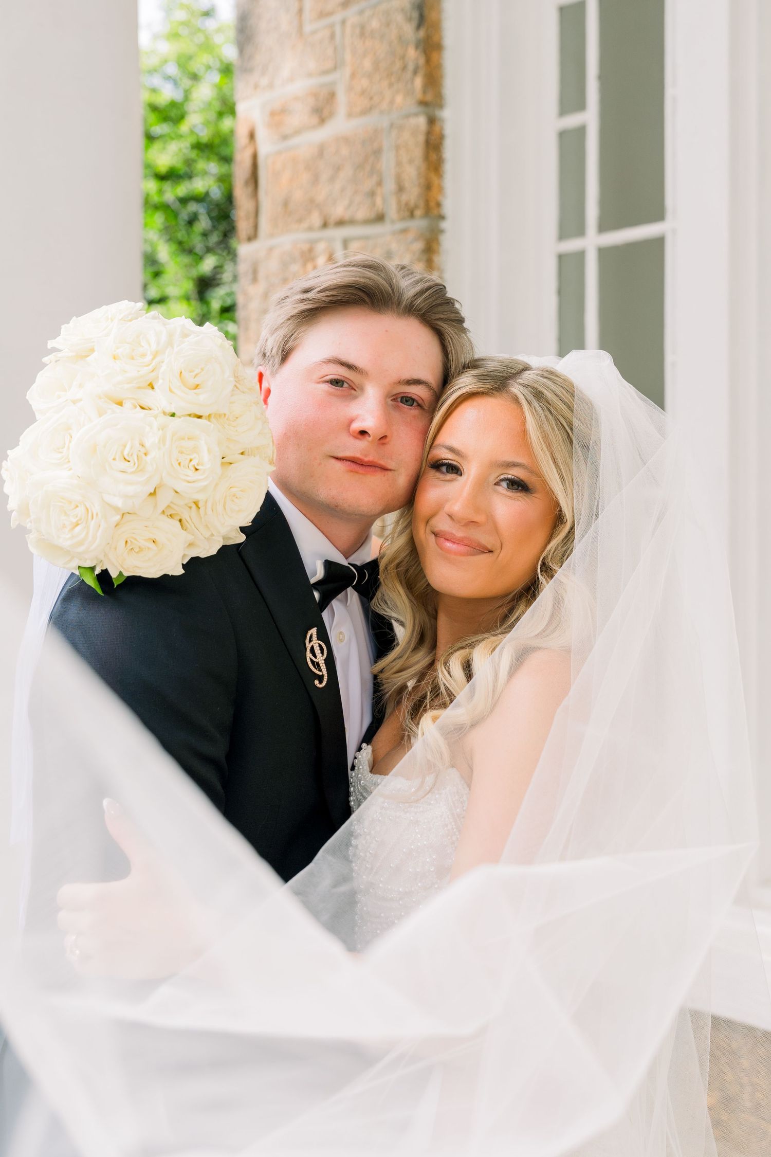 A bride and groom pose together, she holds white roses bouquet, veil flowing, both smiling softly at camera.