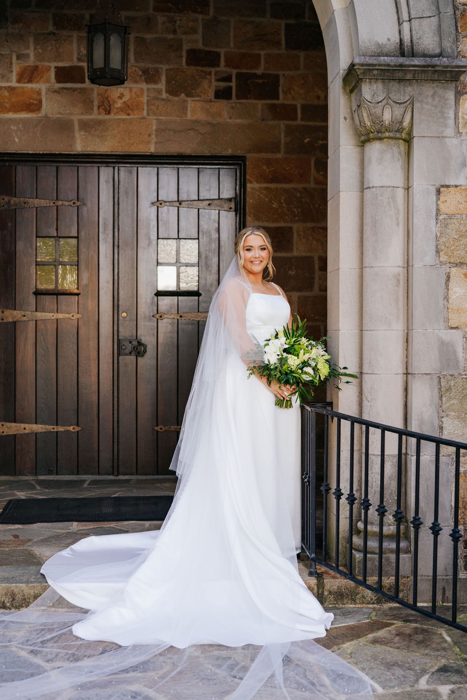 Wedding dress with long train photographed against stone church entrance with bouquet of white and green flowers.