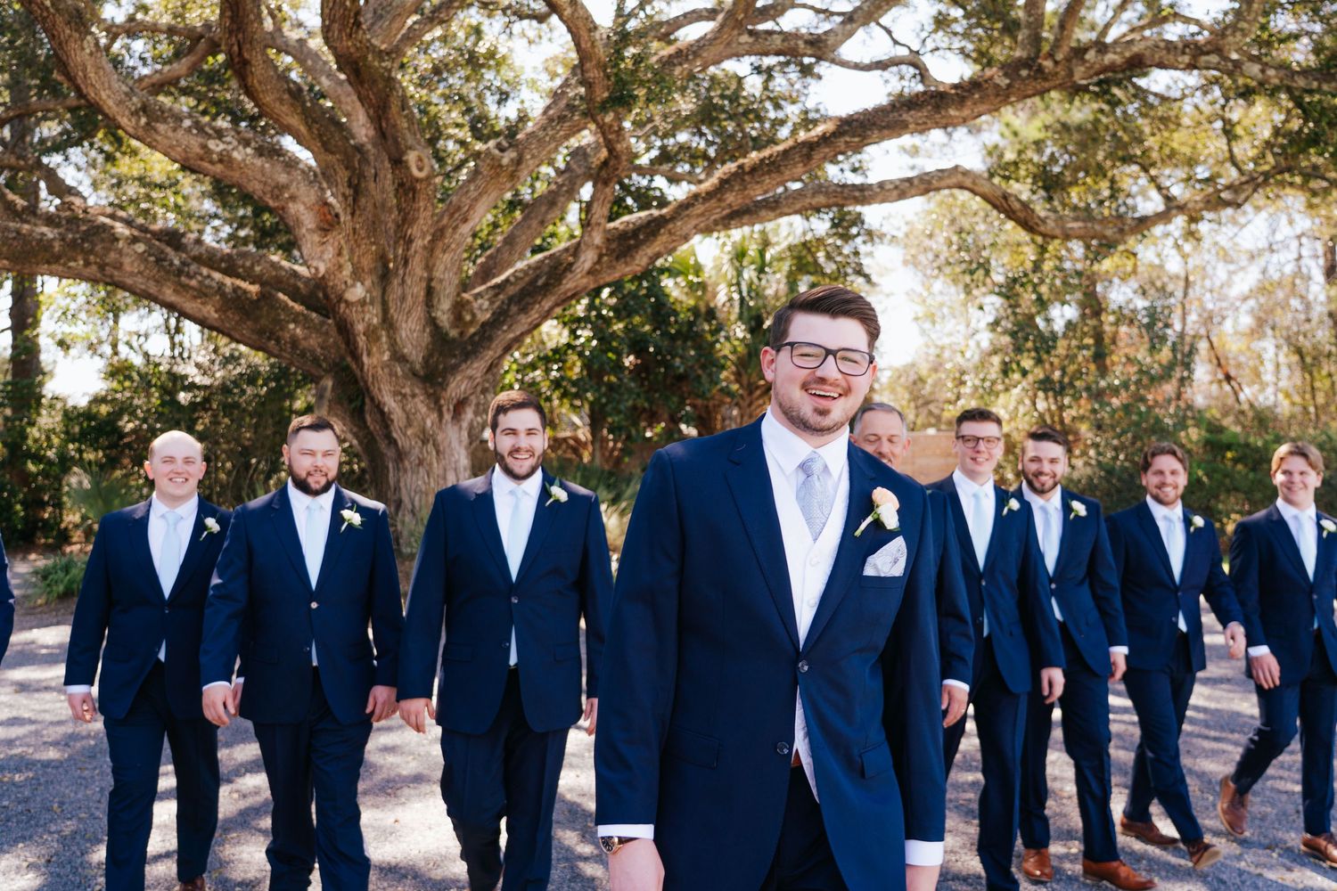 Groomsmen in navy suits pose together under large oak trees for wedding photos.
