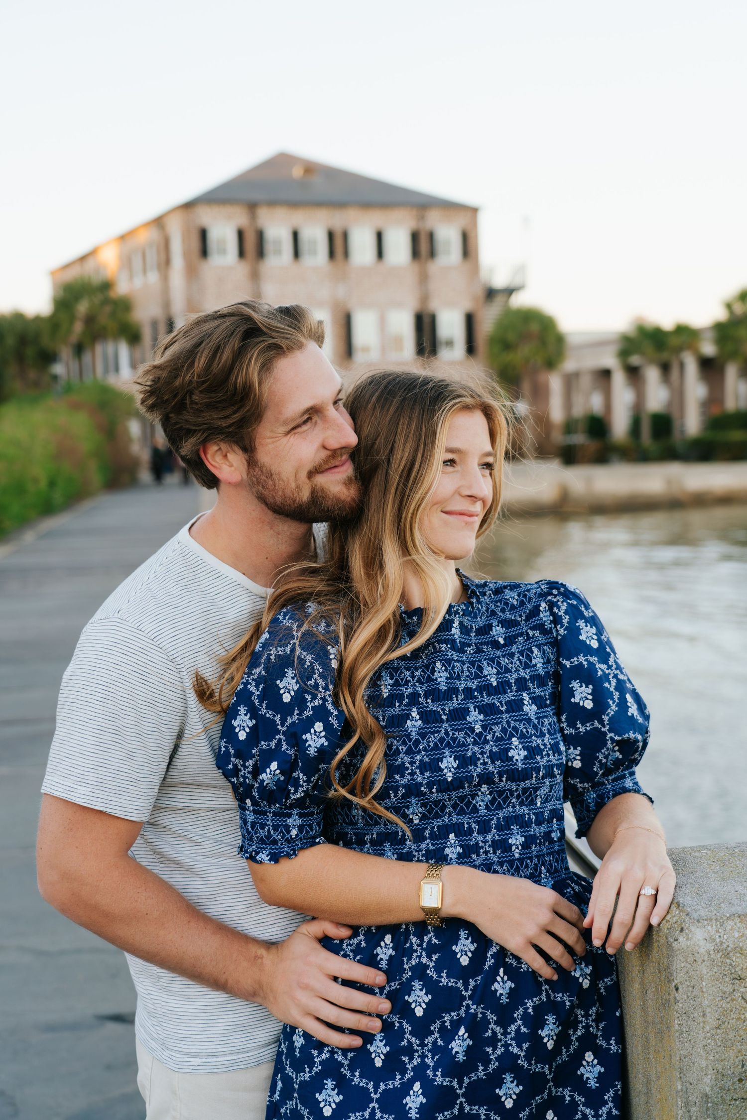 A couple embraces by the waterfront with historic buildings in the background.