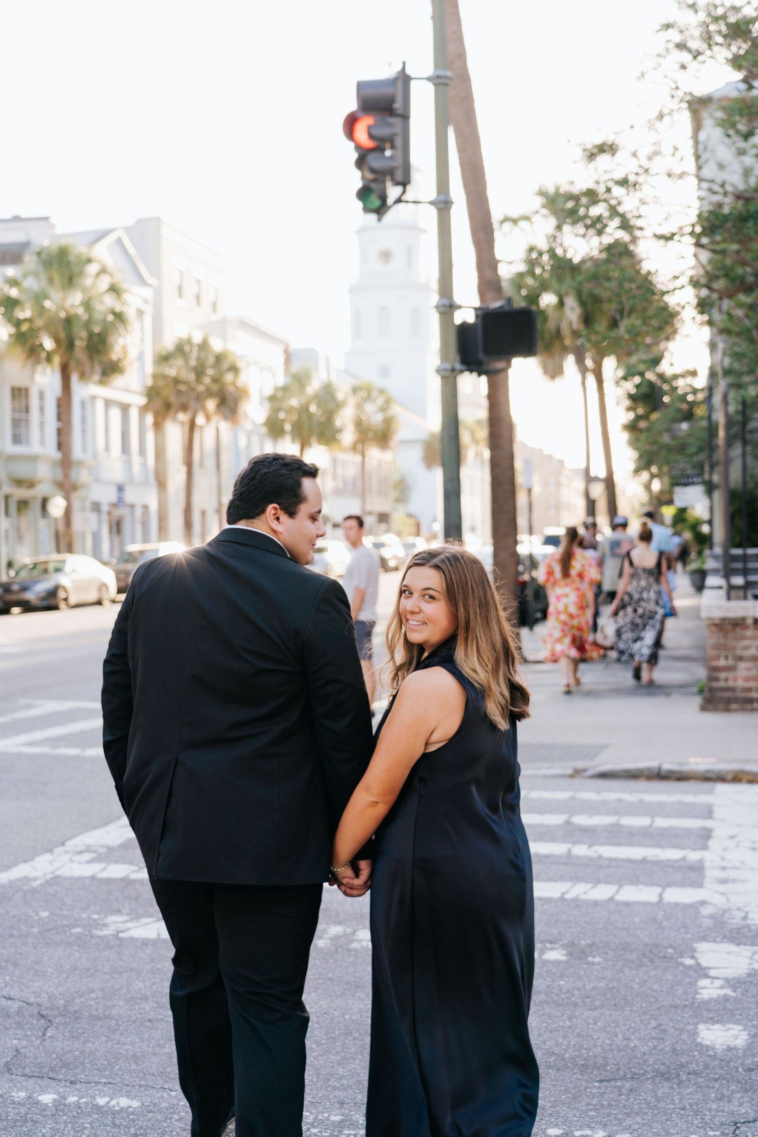 Couple in formal attire walks along a palm-lined city street at sunset.