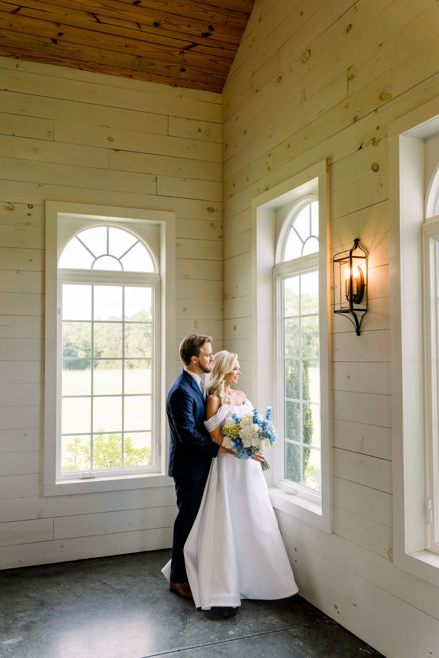 Bride and groom share a romantic moment in a bright chapel with arched windows, white shiplap walls, and a blue bridal bouquet.