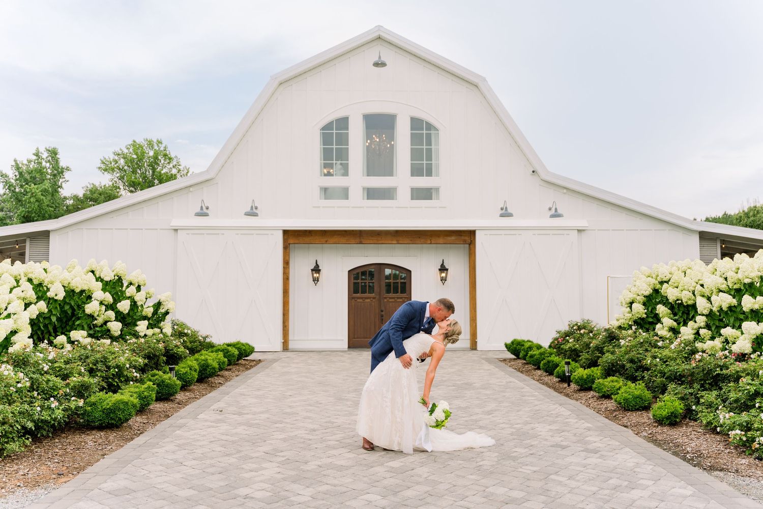 Bride and groom sharing a romantic dip kiss in front of a white barn wedding venue surrounded by hydrangeas.