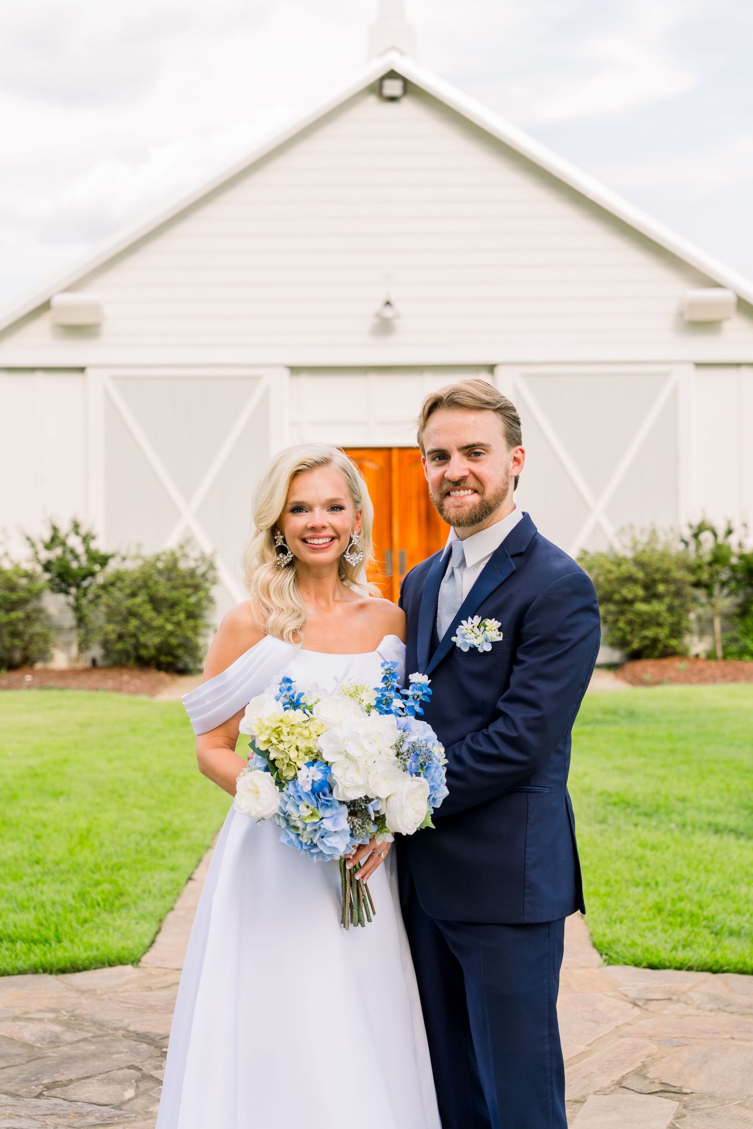 Smiling bride and groom posing before white barn venue, bride in off-shoulder gown with blue hydrangea bouquet.