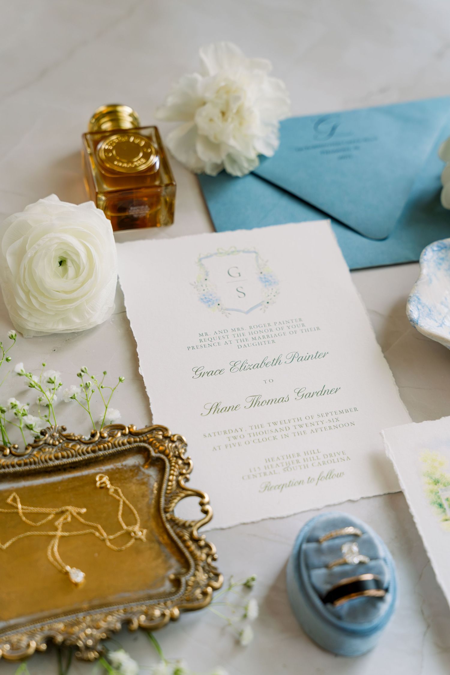 Elegant wedding invitation suite with blue envelope, rings, flowers, and gold tray on marble surface.