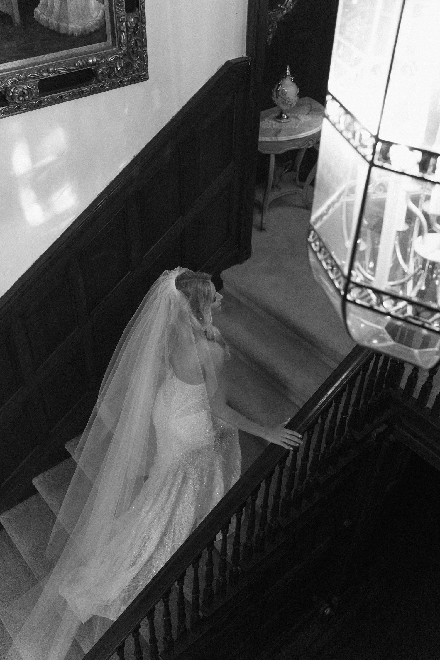 Black and white photo of a bride in a wedding gown and veil descending an elegant staircase in a grand historic venue.