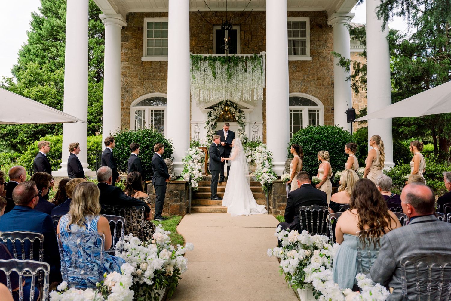 Outdoor wedding ceremony at elegant brick estate with white columns, floral aisle decor, and guests seated on both sides.