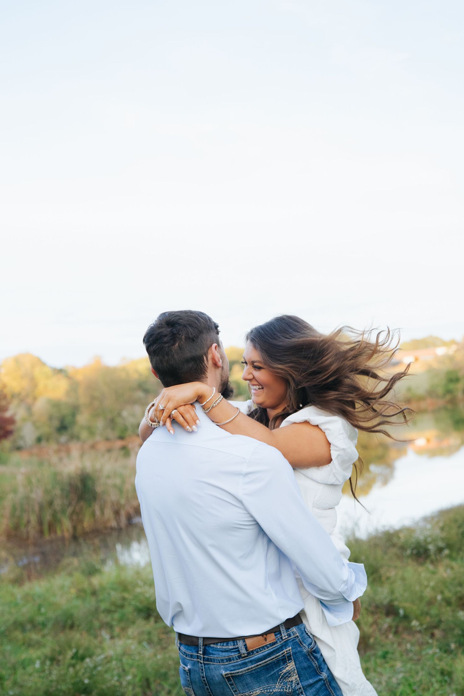 A couple in casual attire spins joyfully together in a grassy field at sunset near a lake.