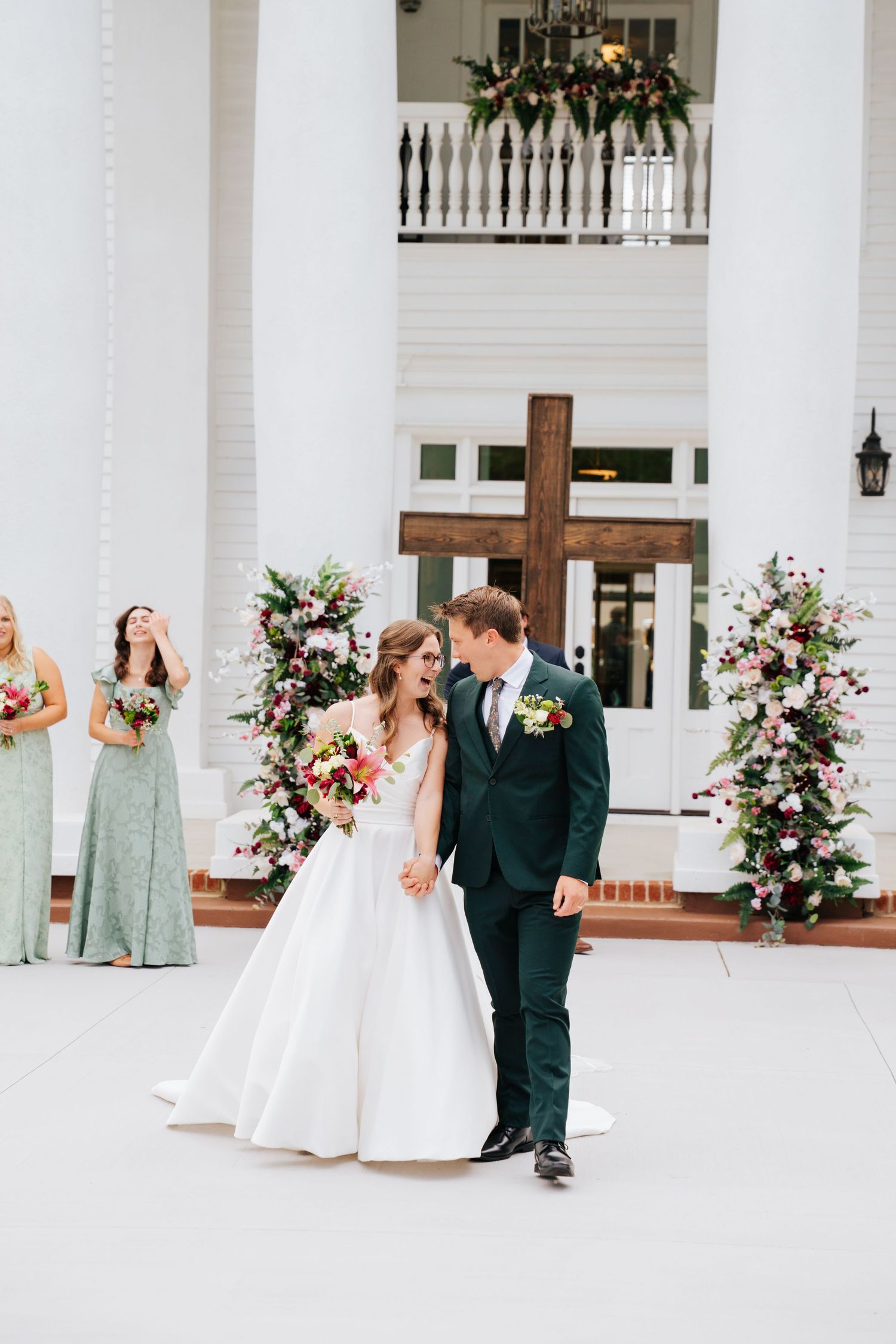 Wedding party celebrates with joyful laughter outside a white columned venue decorated with pink floral arrangements.