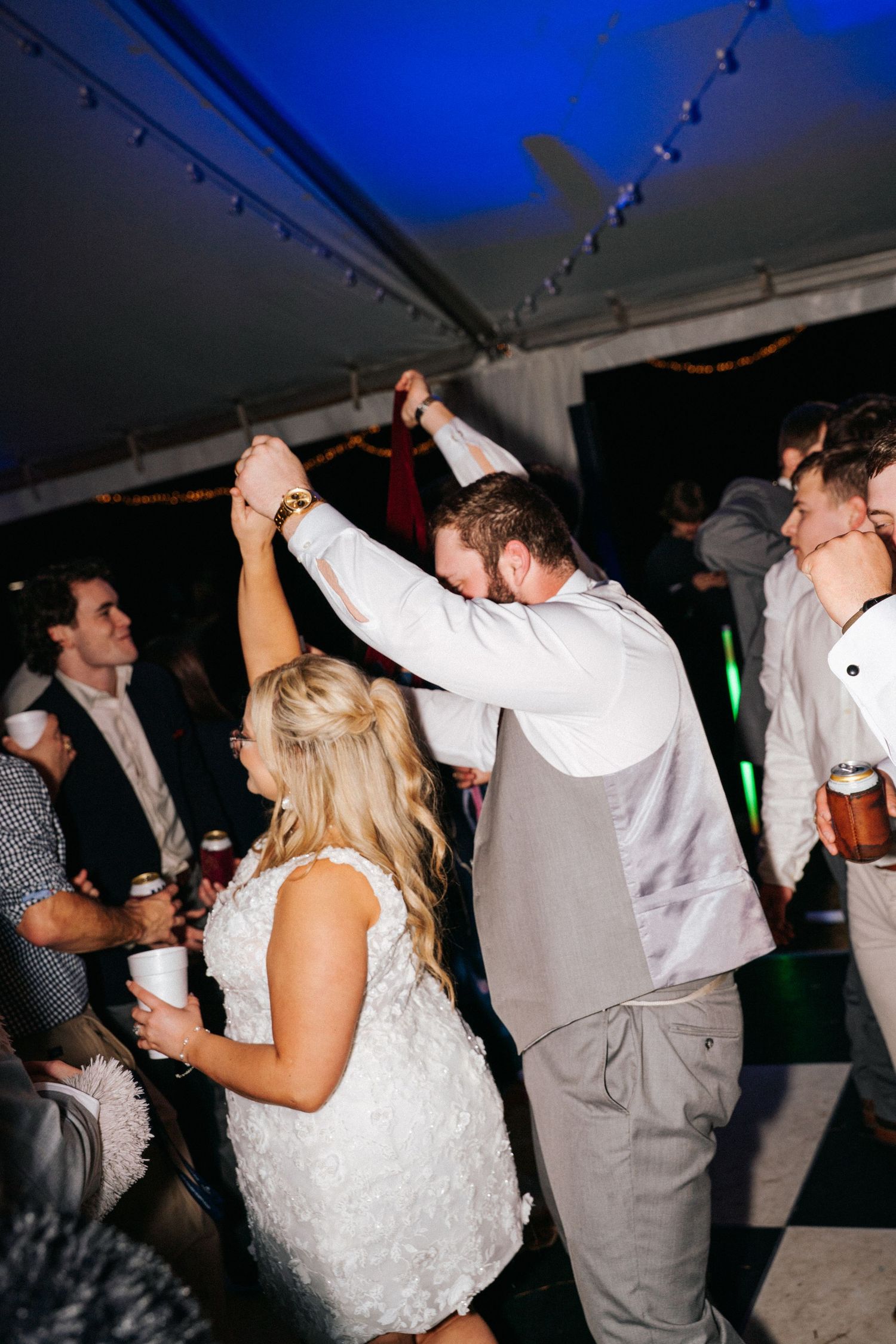 Wedding guests dance and celebrate on a dark dance floor illuminated by blue lighting at an evening reception.
