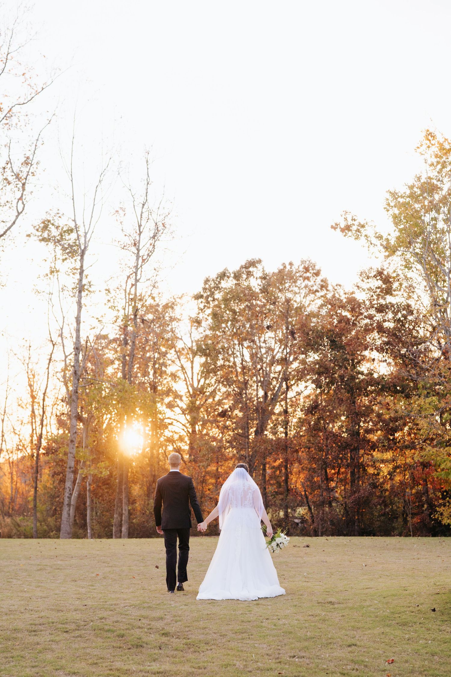 Newlyweds walk hand in hand across grassy field during golden hour sunset.