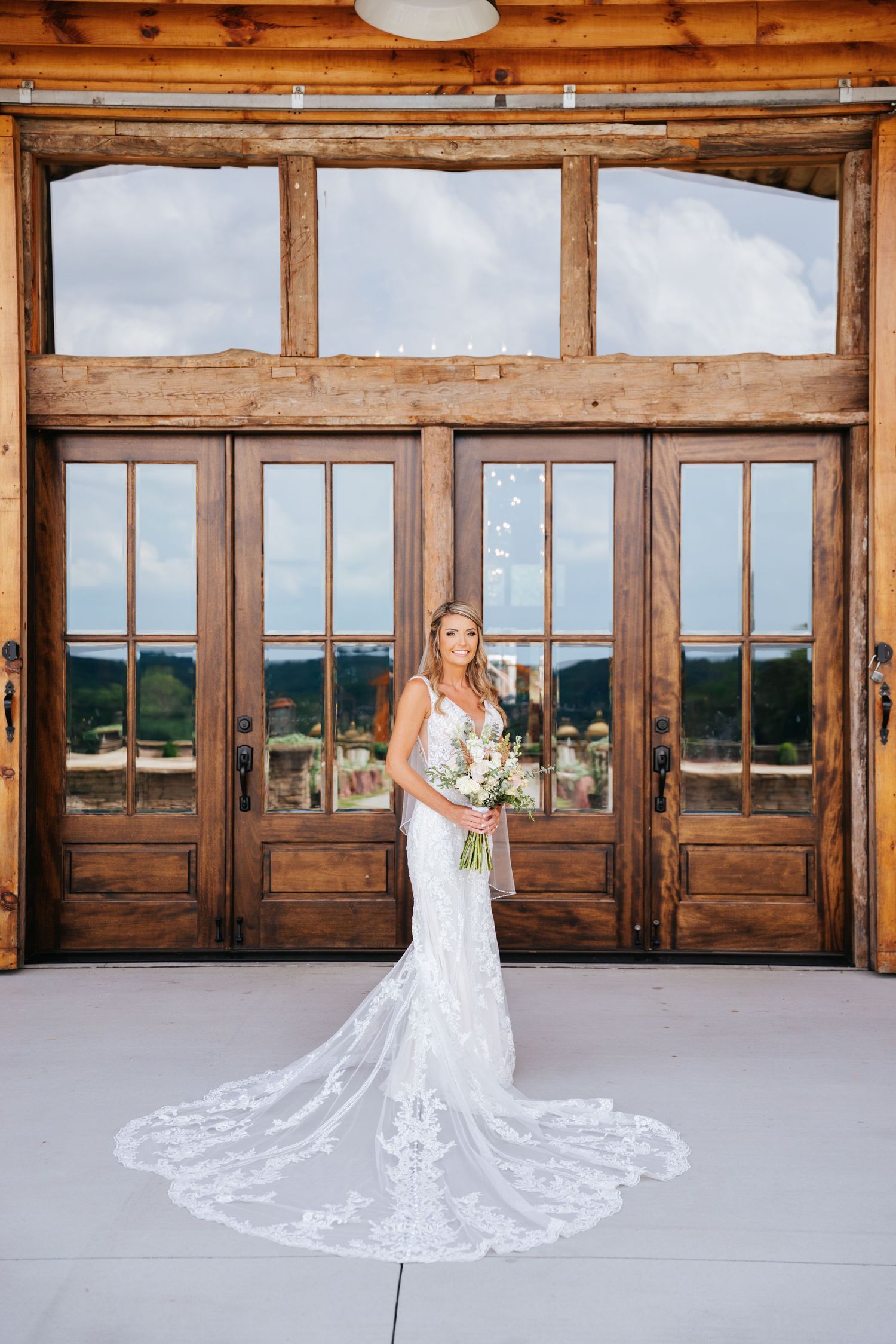 Bride in lace wedding gown with long train poses in rustic wooden venue.
