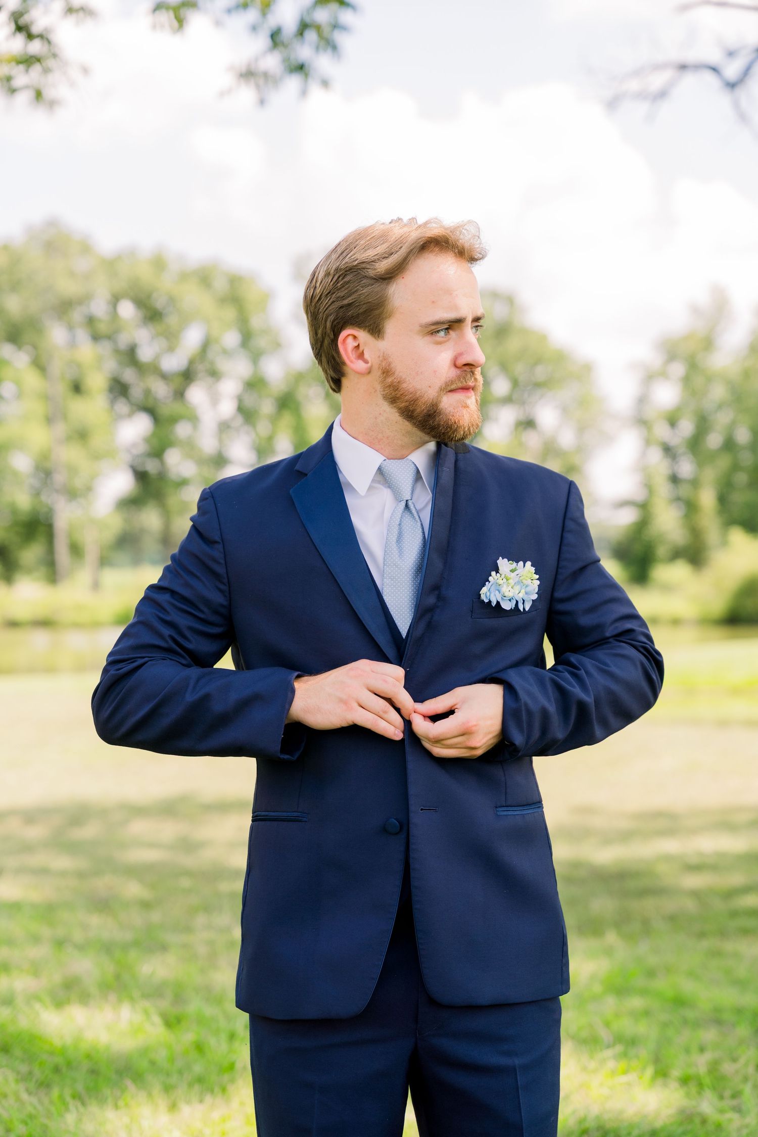 A dapper groom in a navy blue suit with boutonniere buttons his jacket outdoors in a lush green park setting.
