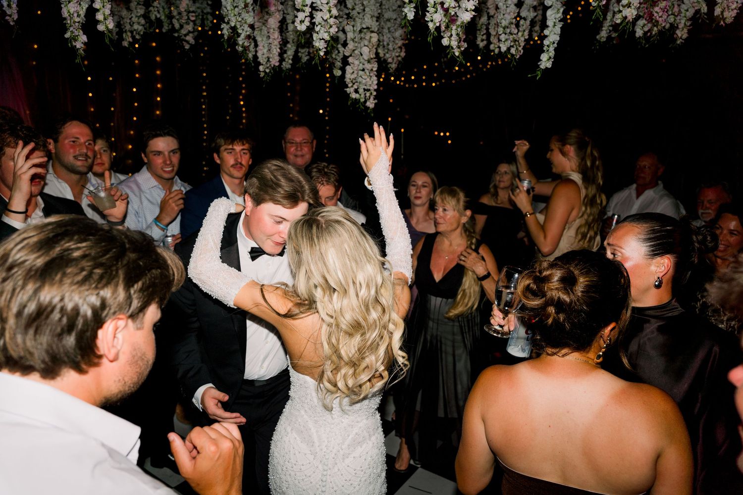 Bride and groom share a joyful dance surrounded by celebrating wedding guests at a festive evening reception.