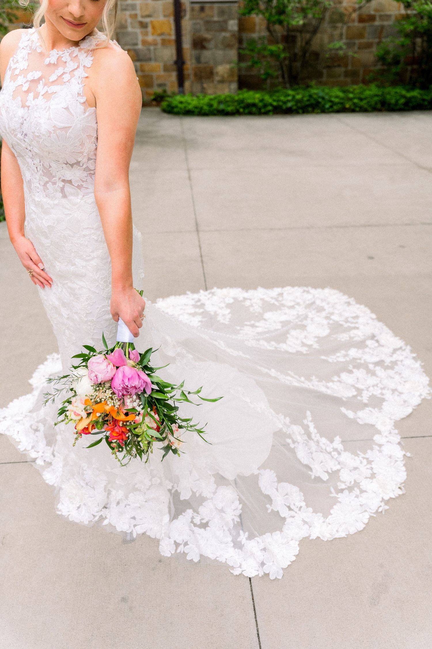 Bride in lace mermaid wedding dress holding colorful bouquet with flowing floral veil train on concrete.
