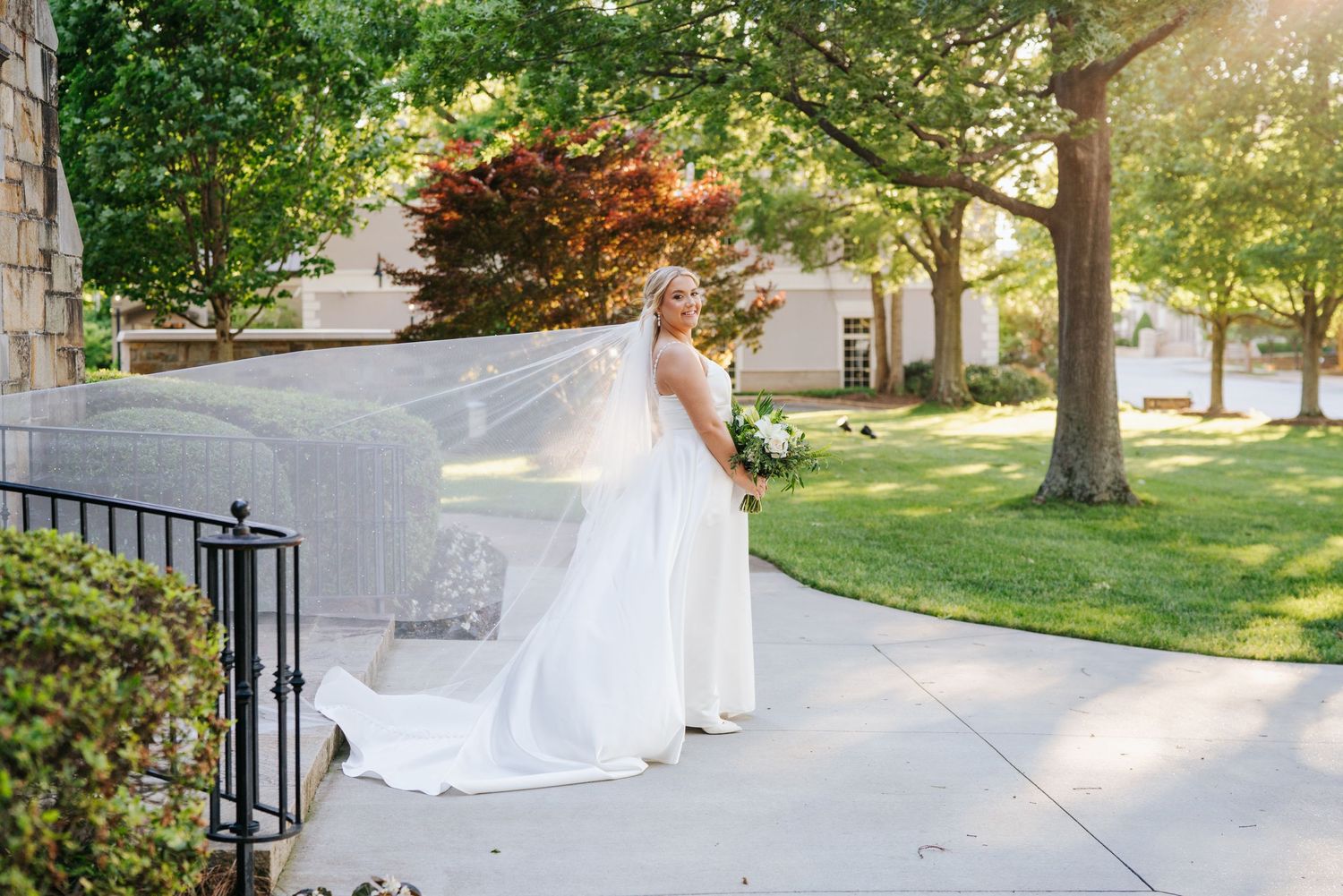 Sunlit wedding portrait with flowing veil and dress in outdoor garden setting.