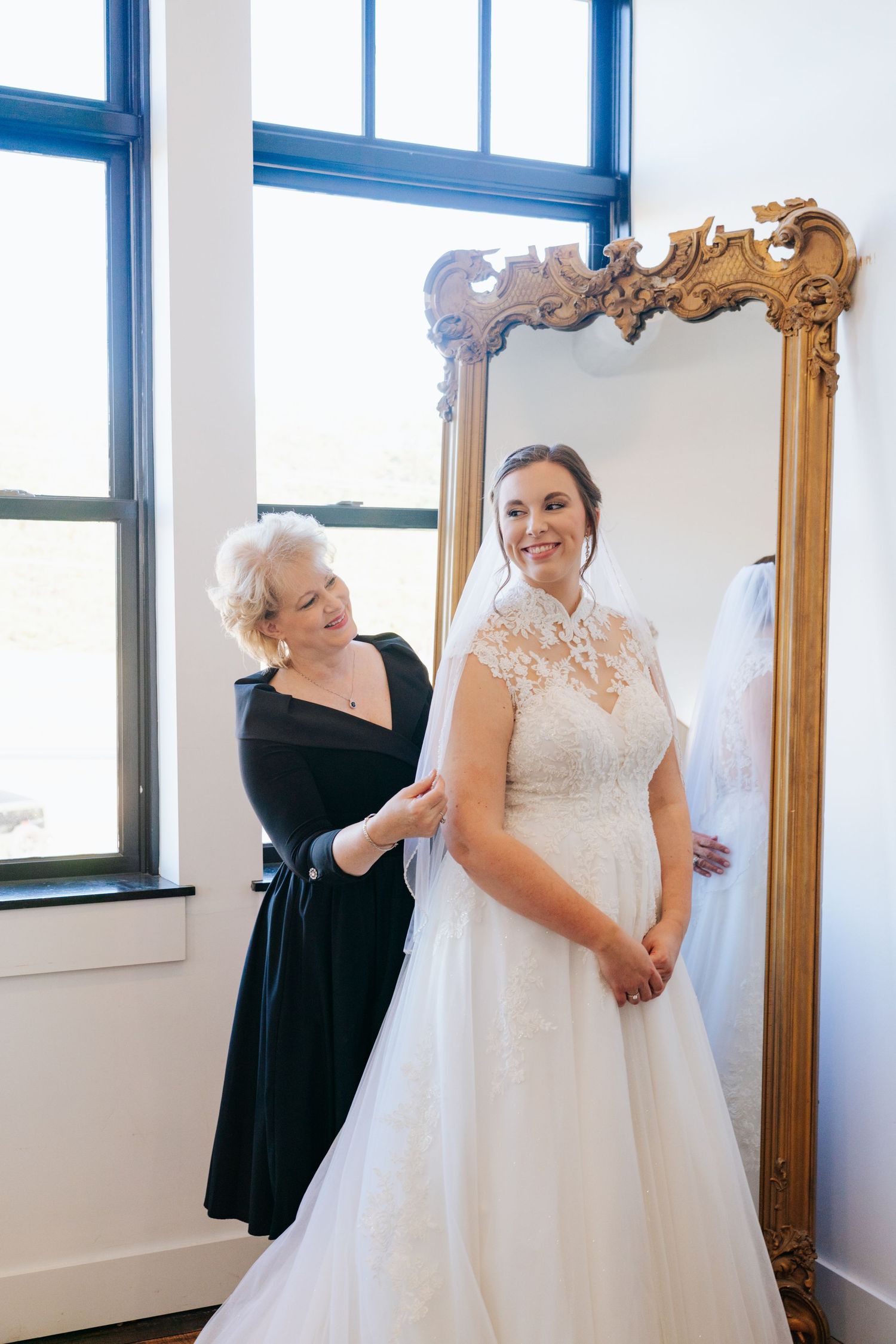 Bride trying on her white lace wedding gown in front of an ornate gold mirror.