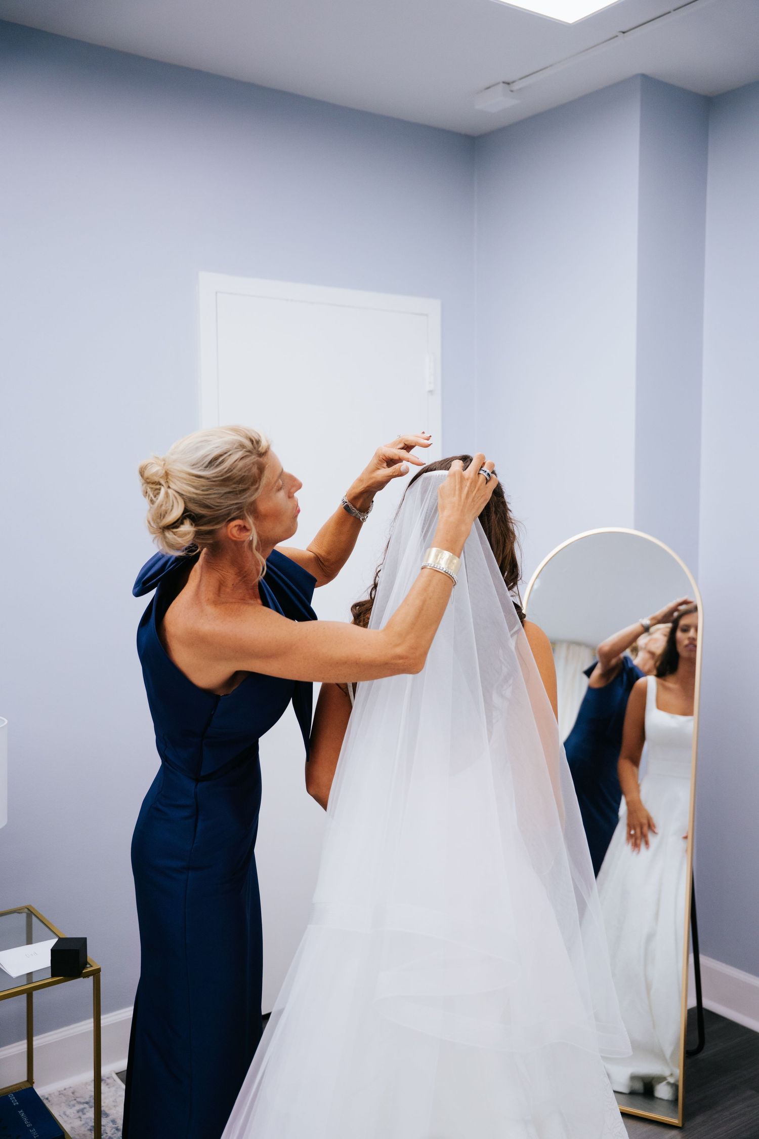Mother adjusts a white wedding veil in a bright room with blue walls.