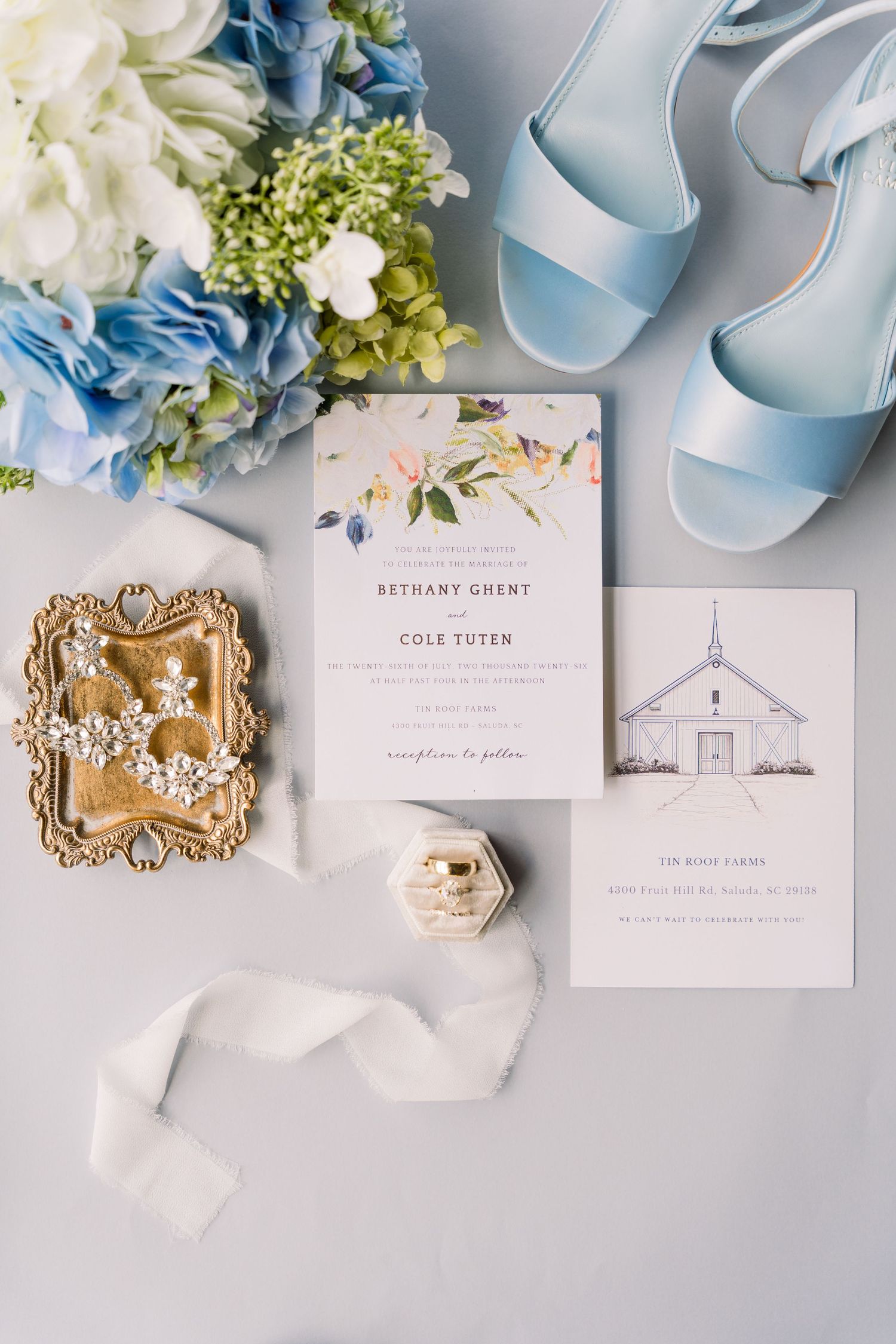 Elegant wedding flat lay with floral invitation, light blue heels, hydrangea bouquet, diamond ring box, and gold jewelry tray.