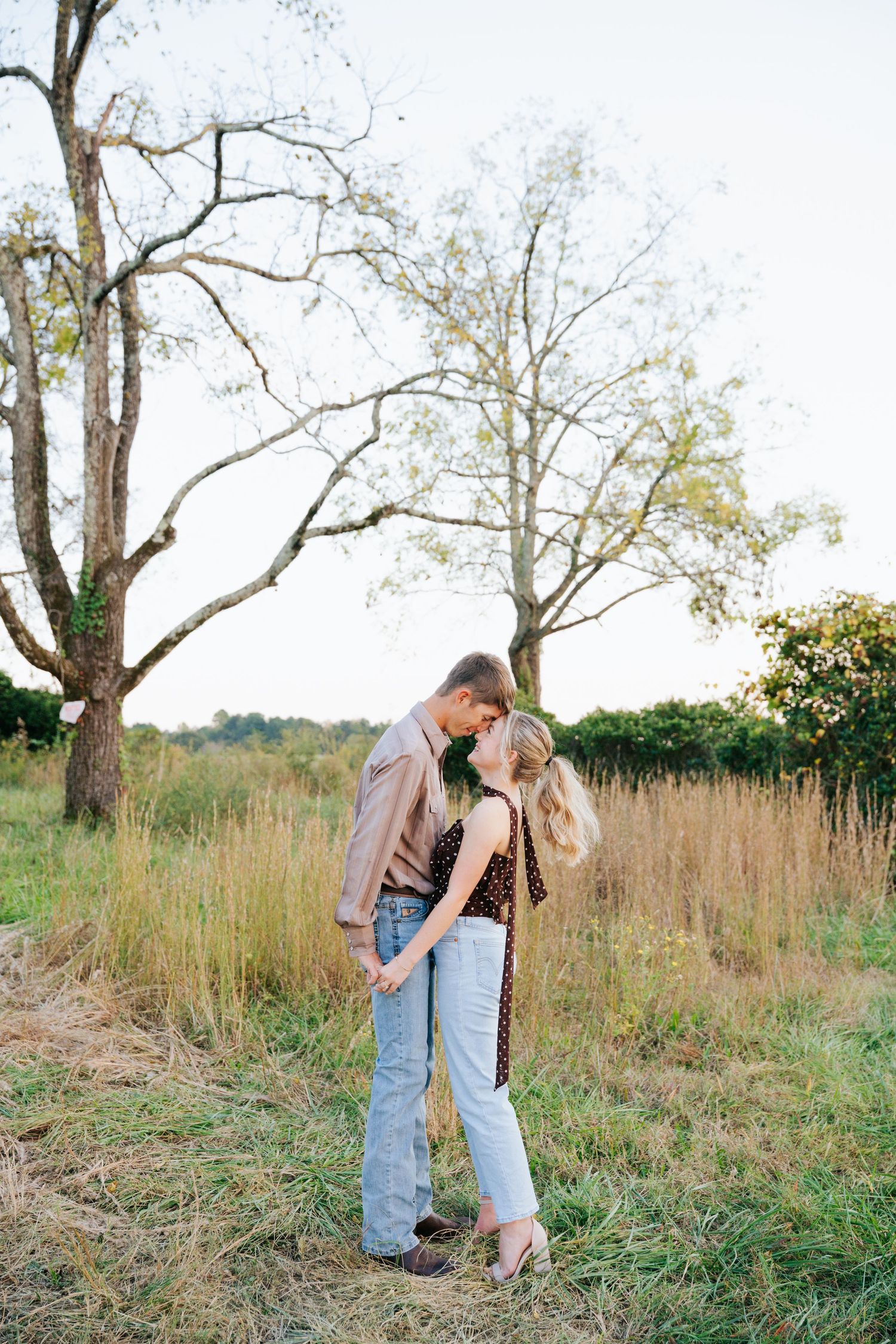A couple shares a romantic moment together in a serene field with tall grass and bare trees in the background.