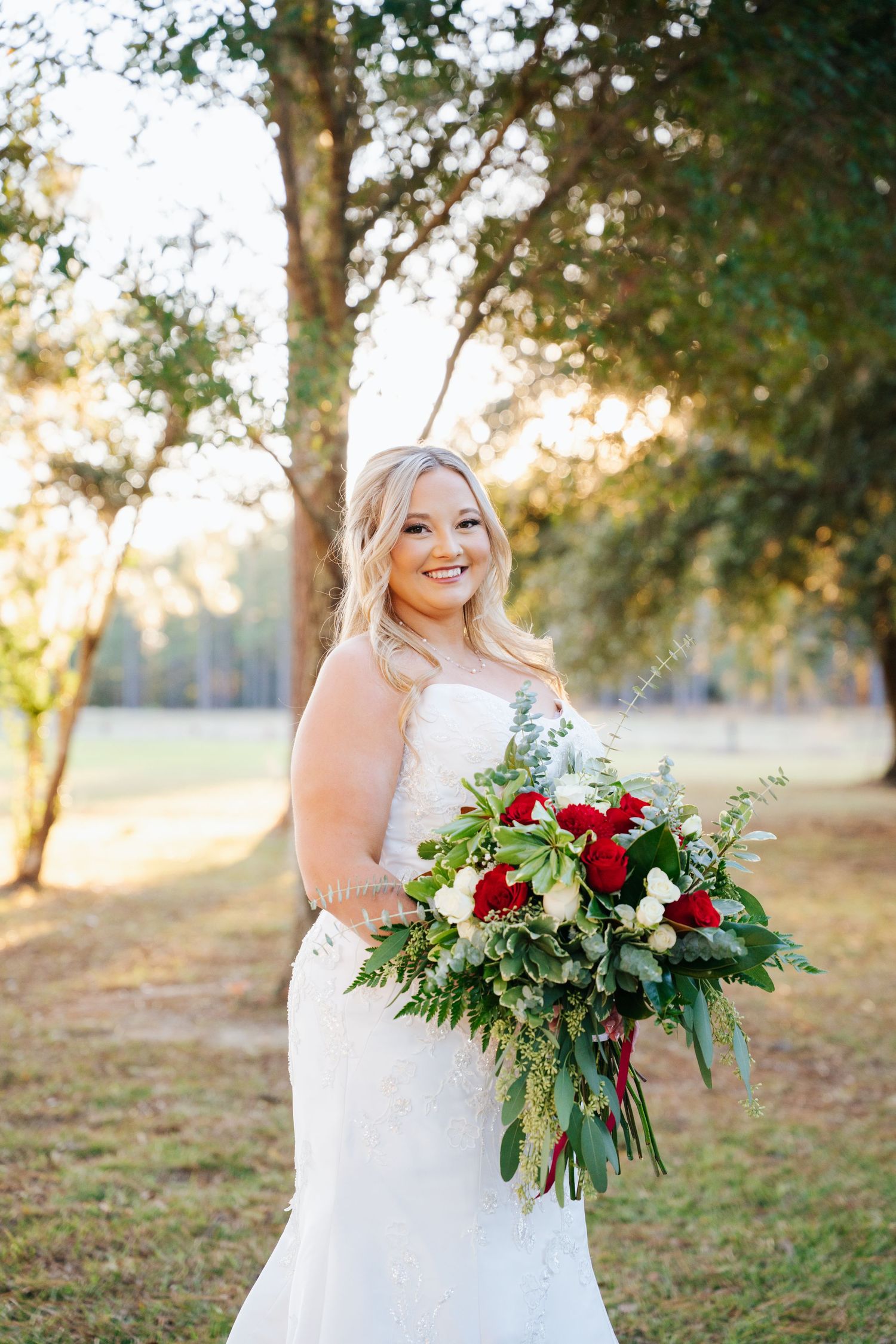 A bride in a white lace dress holds a vibrant red and green bouquet while posing outdoors during golden hour.