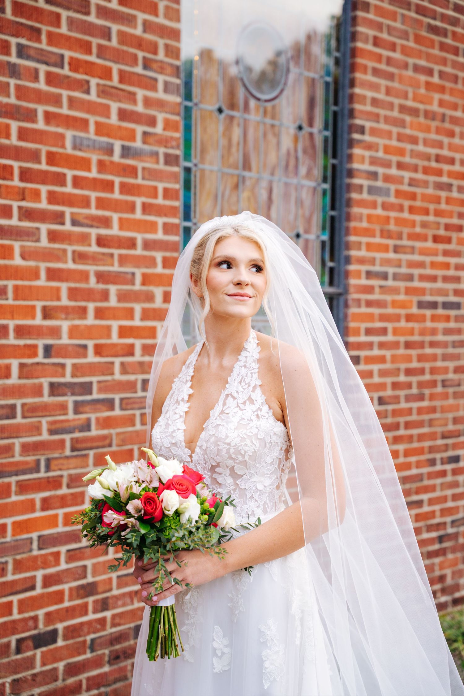 A bride in a white lace gown with a veil poses against a brick wall while holding a pink and red floral bouquet.