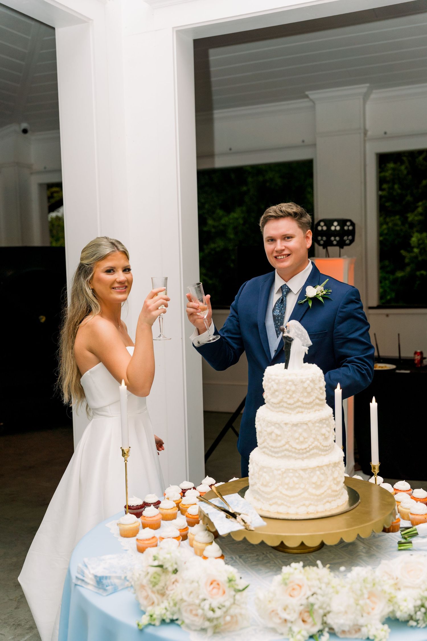 Bride and groom toasting with champagne glasses beside a white three-tier wedding cake at their reception.
