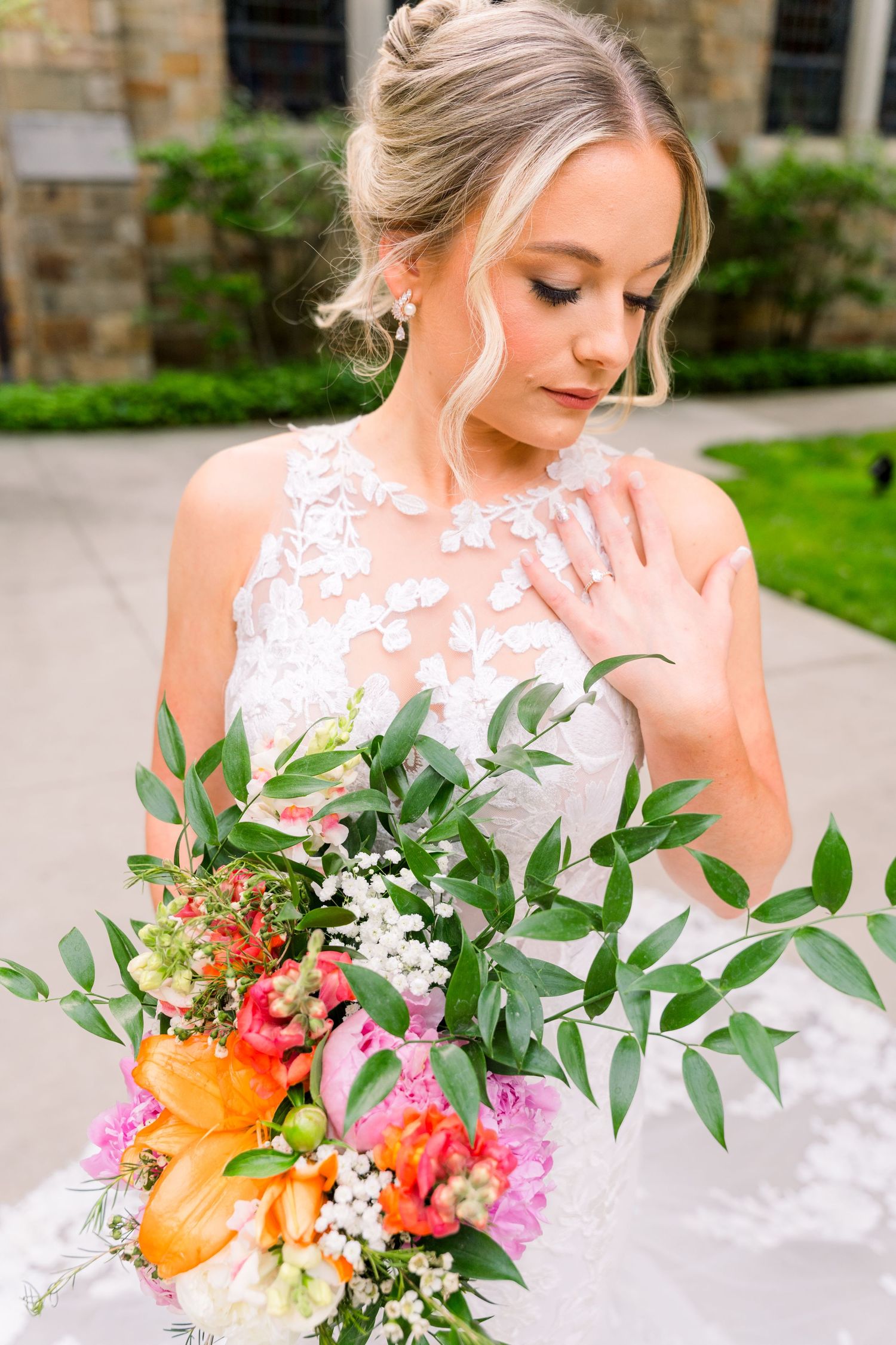 Bride in lace wedding gown holds vibrant colorful bouquet with greenery, gazing down elegantly outdoors.