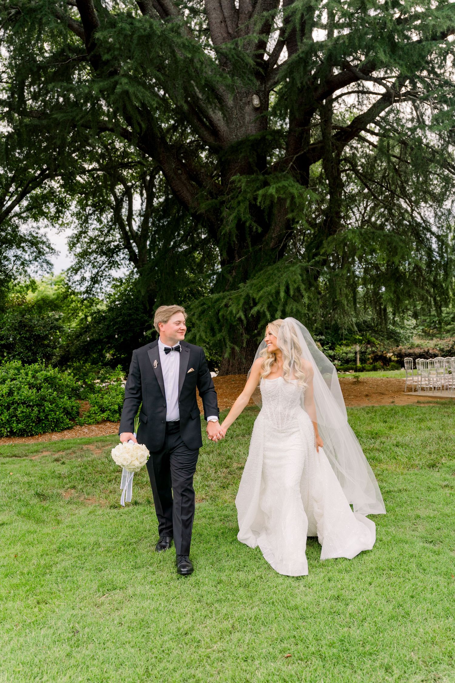 A bride and groom walk hand-in-hand on green grass beneath a large tree, she in a white gown with veil, he in a black tuxedo.