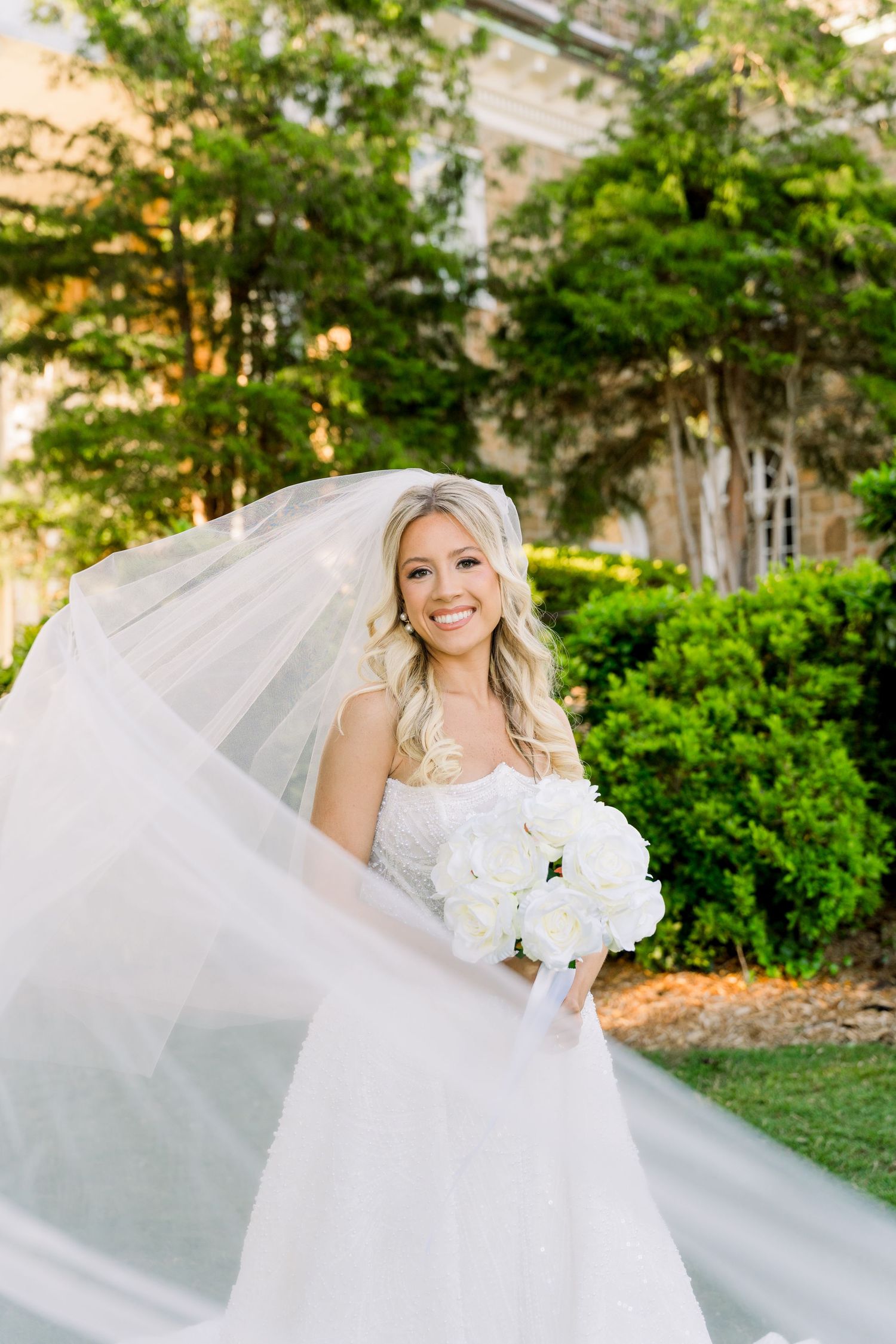 Smiling bride in white gown holds bouquet of white roses, veil flowing in breeze, posing outdoors in lush garden setting.