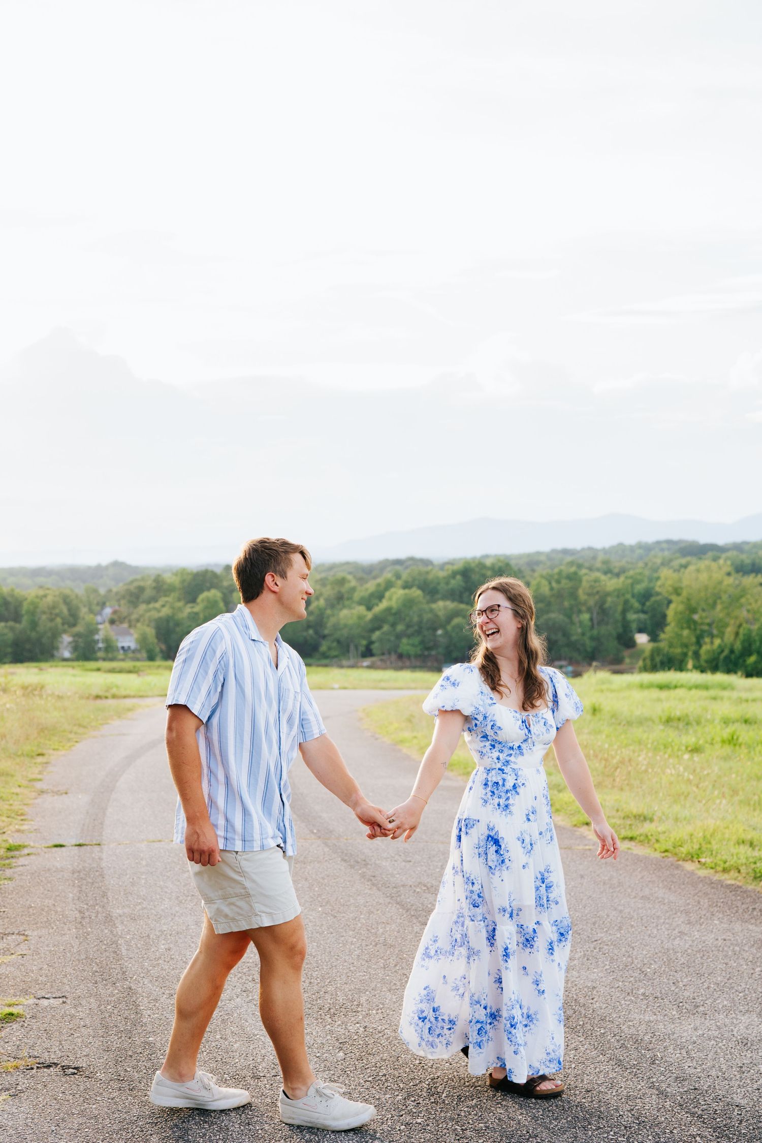 A couple walks hand in hand on a rural road surrounded by green fields and mountains in the distance.