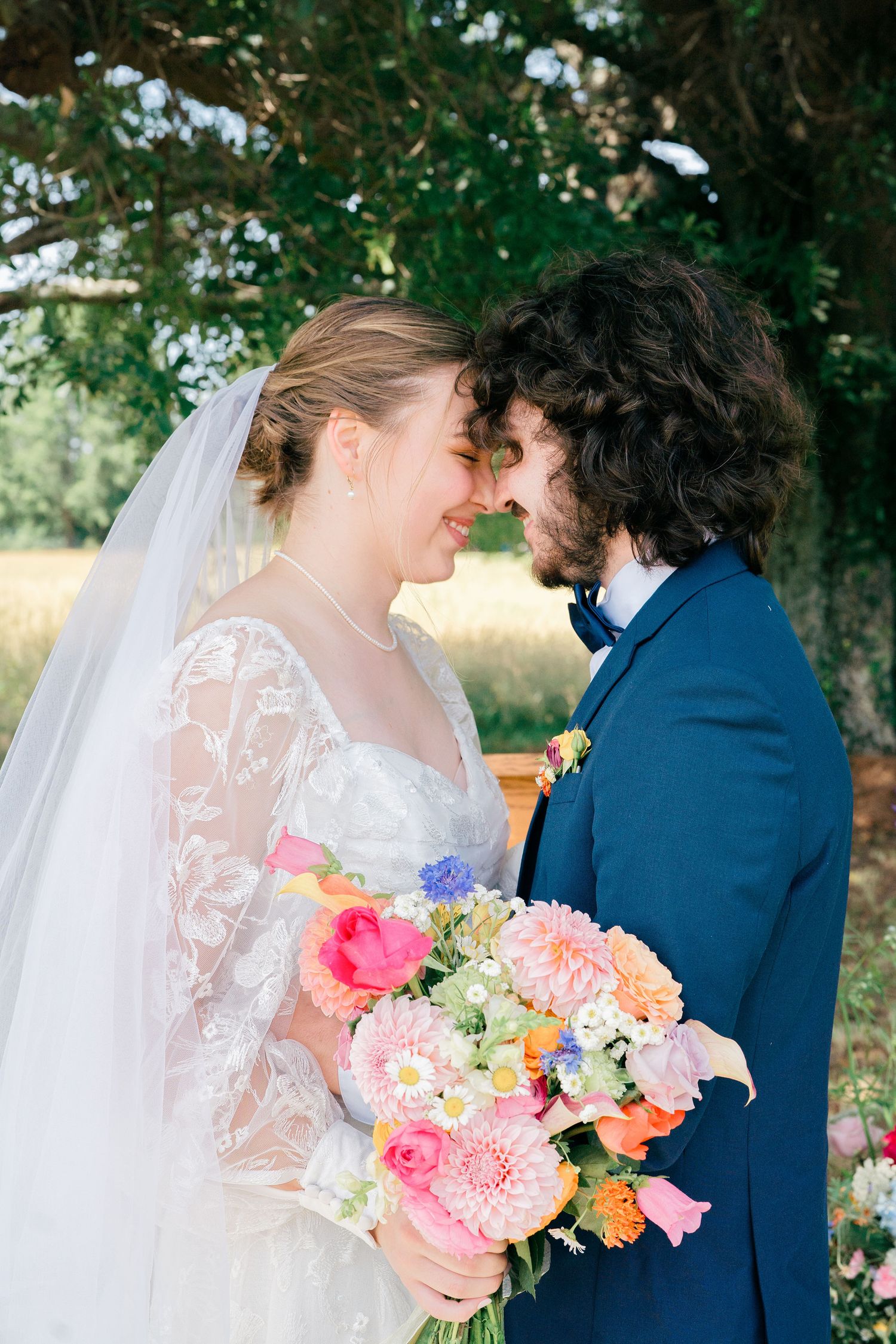 A romantic wedding moment captured with a colorful bouquet of pink and coral flowers against a natural outdoor backdrop.