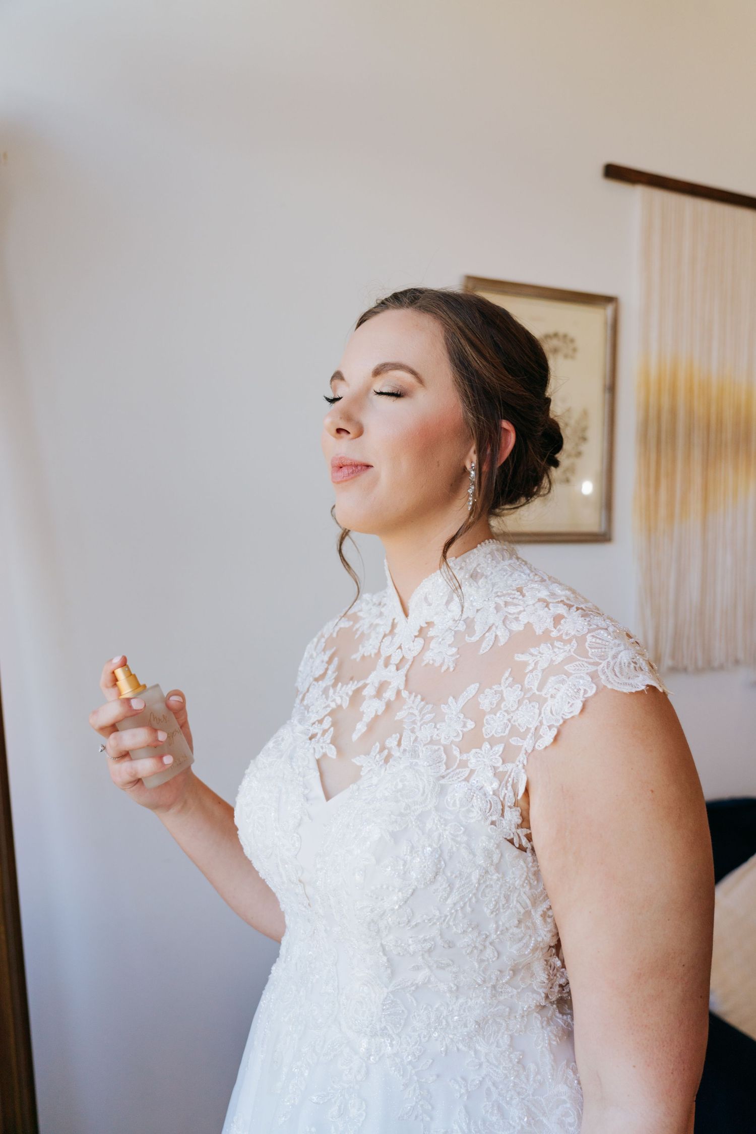 Bride in a lace wedding dress spraying perfume in preparation for the ceremony.