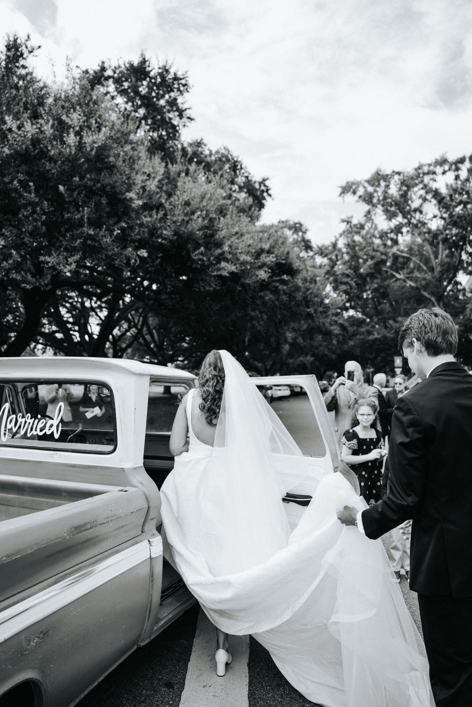 A classic vintage truck helps transport the bride and groom on a tree-lined street.