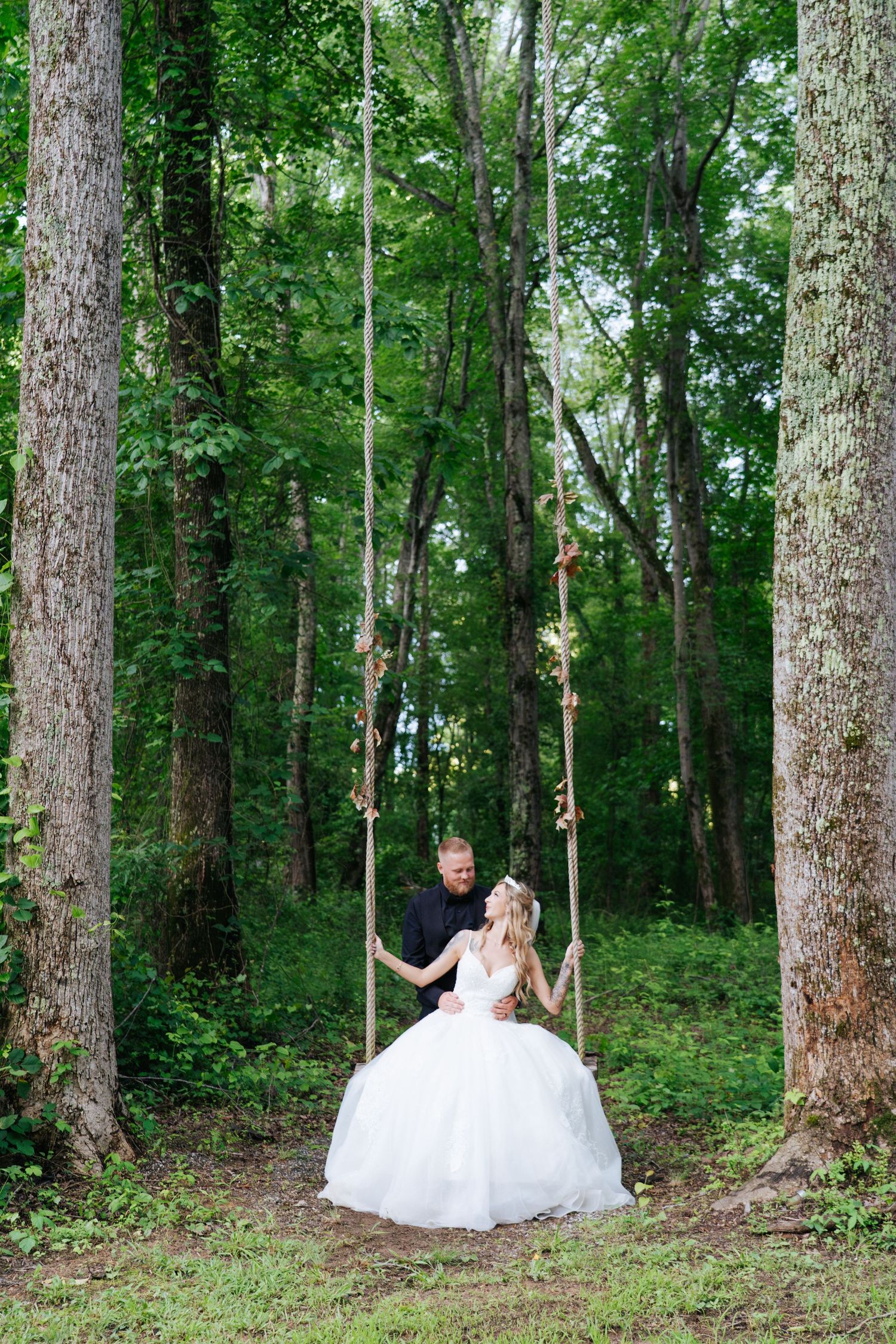 Wedding couple poses between tall trees in a romantic woodland setting.
