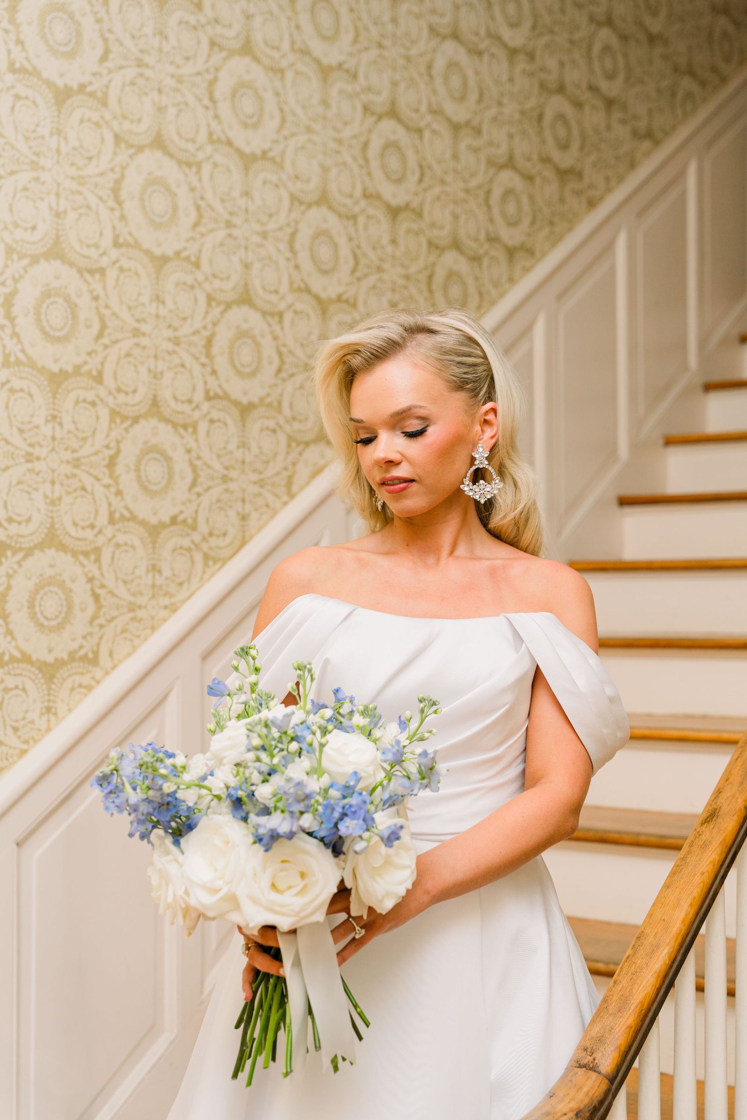 Elegant bride in off-shoulder white gown holding blue and white bouquet on staircase with patterned wallpaper.
