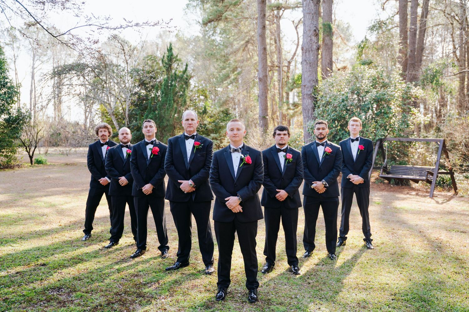 Wedding party in black tuxedos poses together in a scenic outdoor forest setting during a formal celebration.